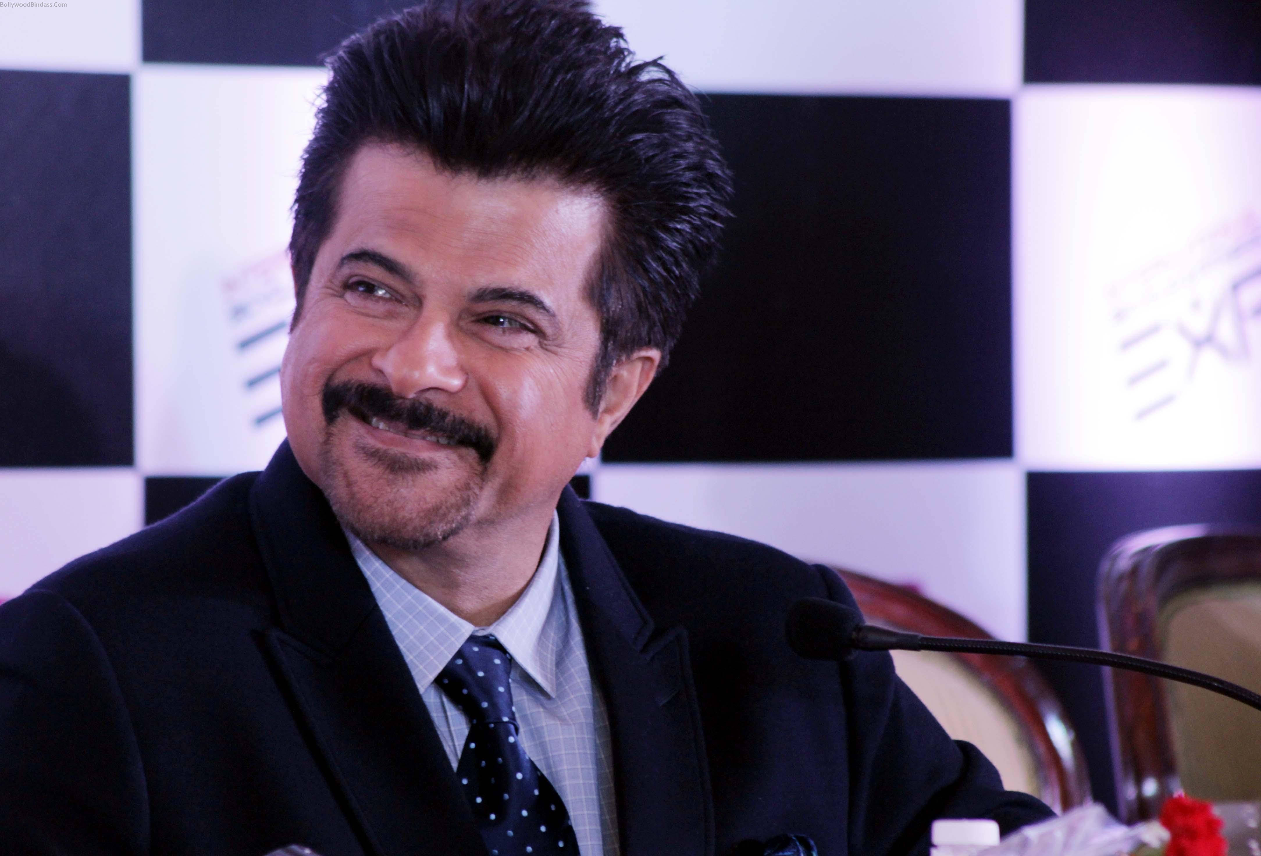 anil kapoor 桌布 壁紙 — 一位留鬍鬚、面帶微笑的男士，身穿深色西裝和波點領帶，坐在麥克風前，背景是黑白格紋圖案。