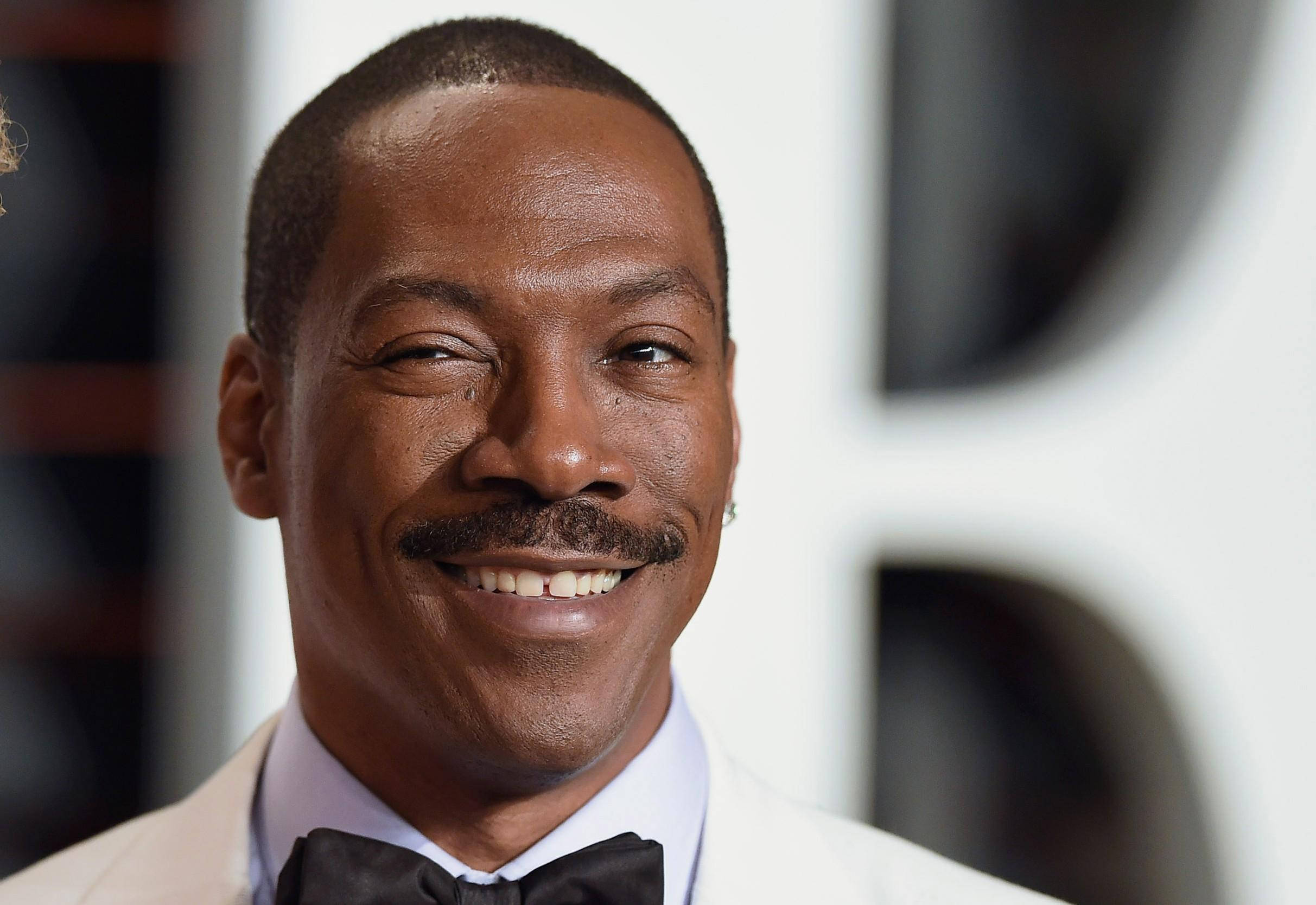 eddie murphy 桌布 壁紙 — 一位留鬍鬚、面帶微笑的男士身穿白色西裝和黑色領結的特寫肖像。
