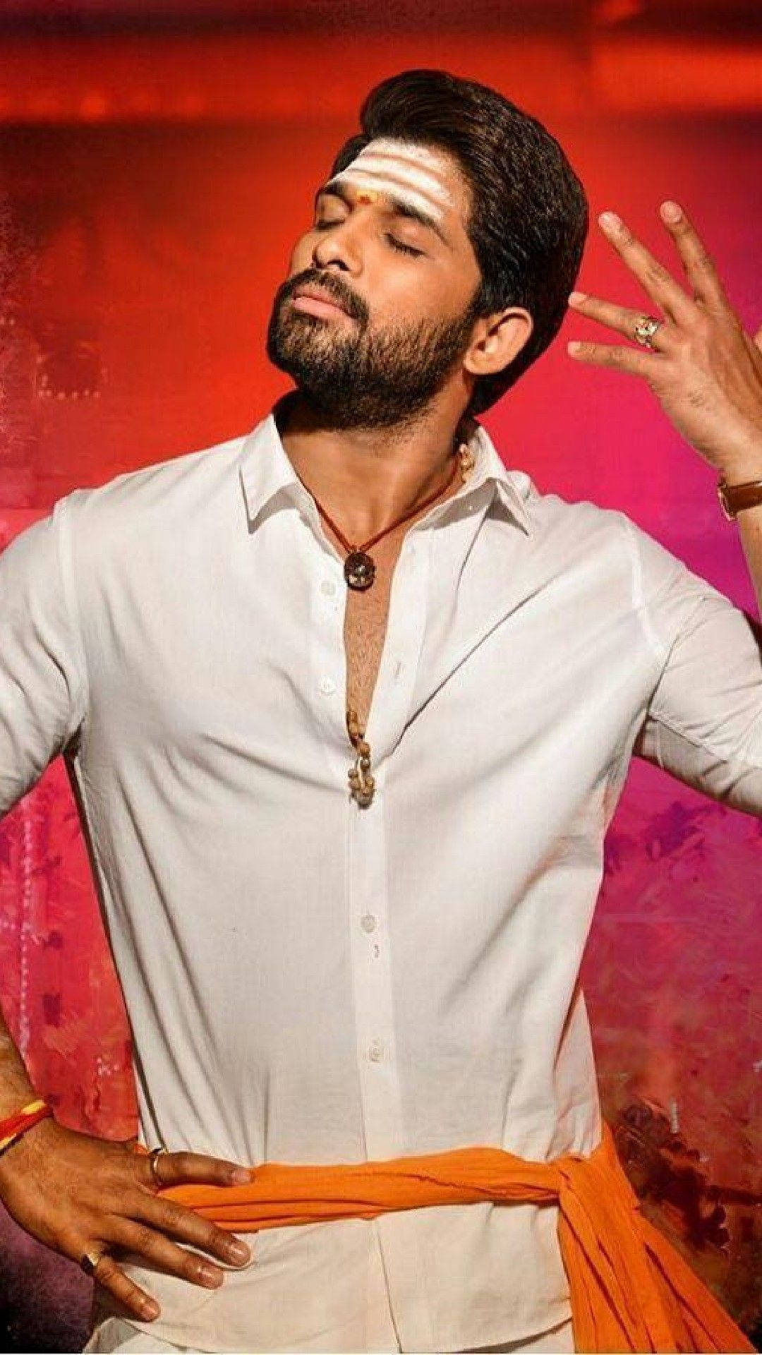 Allu Arjun 桌布 壁紙 — 一位留鬍鬚、額頭有白色標記的男子，自信地穿著白襯衫和橙色腰帶，在鮮豔的紅色和粉紅色背景前擺姿勢。