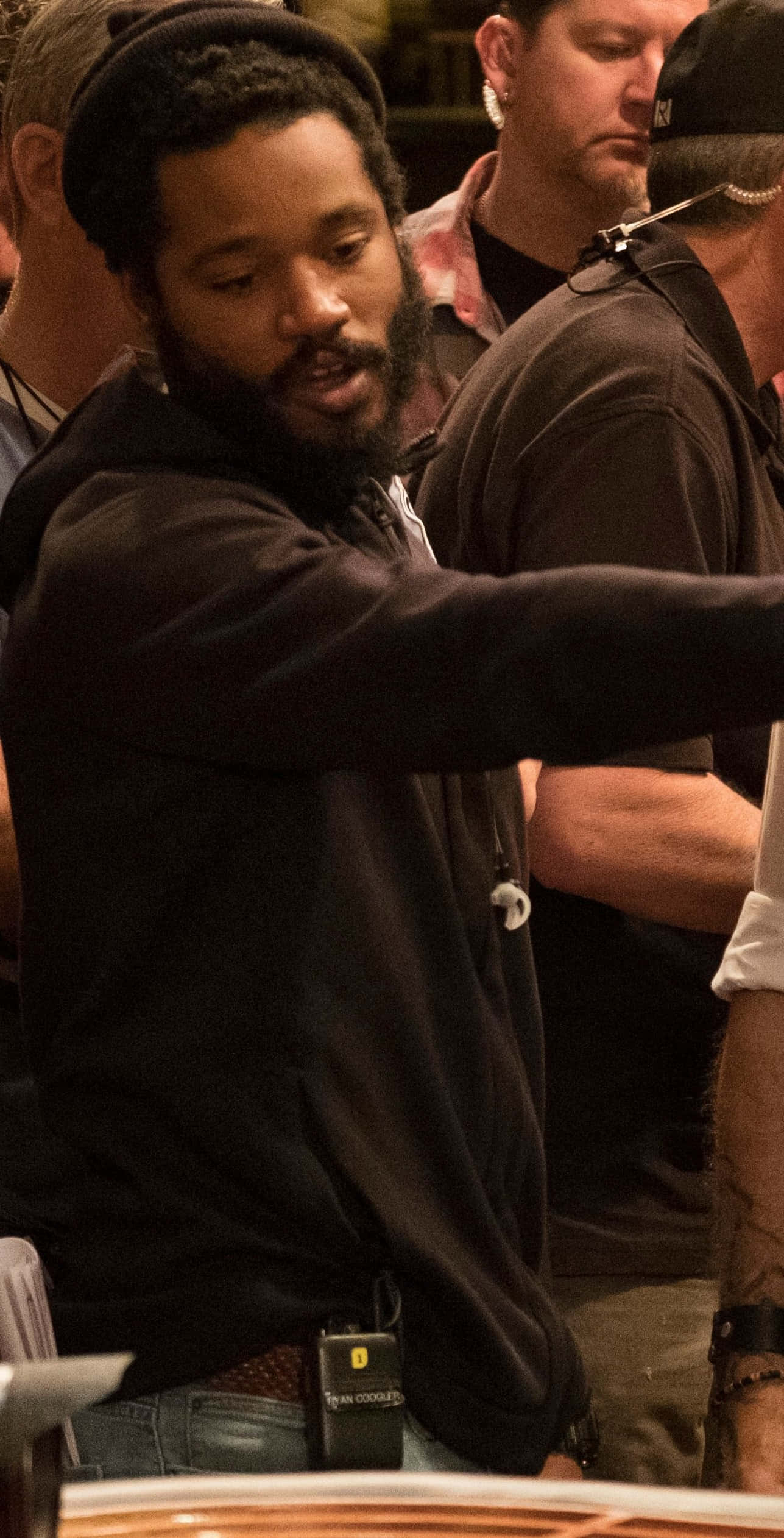 Ryan Coogler 桌布 壁紙 — 一位留鬍鬚戴毛帽的男子在擁擠的室內環境中，在溫暖的燈光下生動地做手勢。