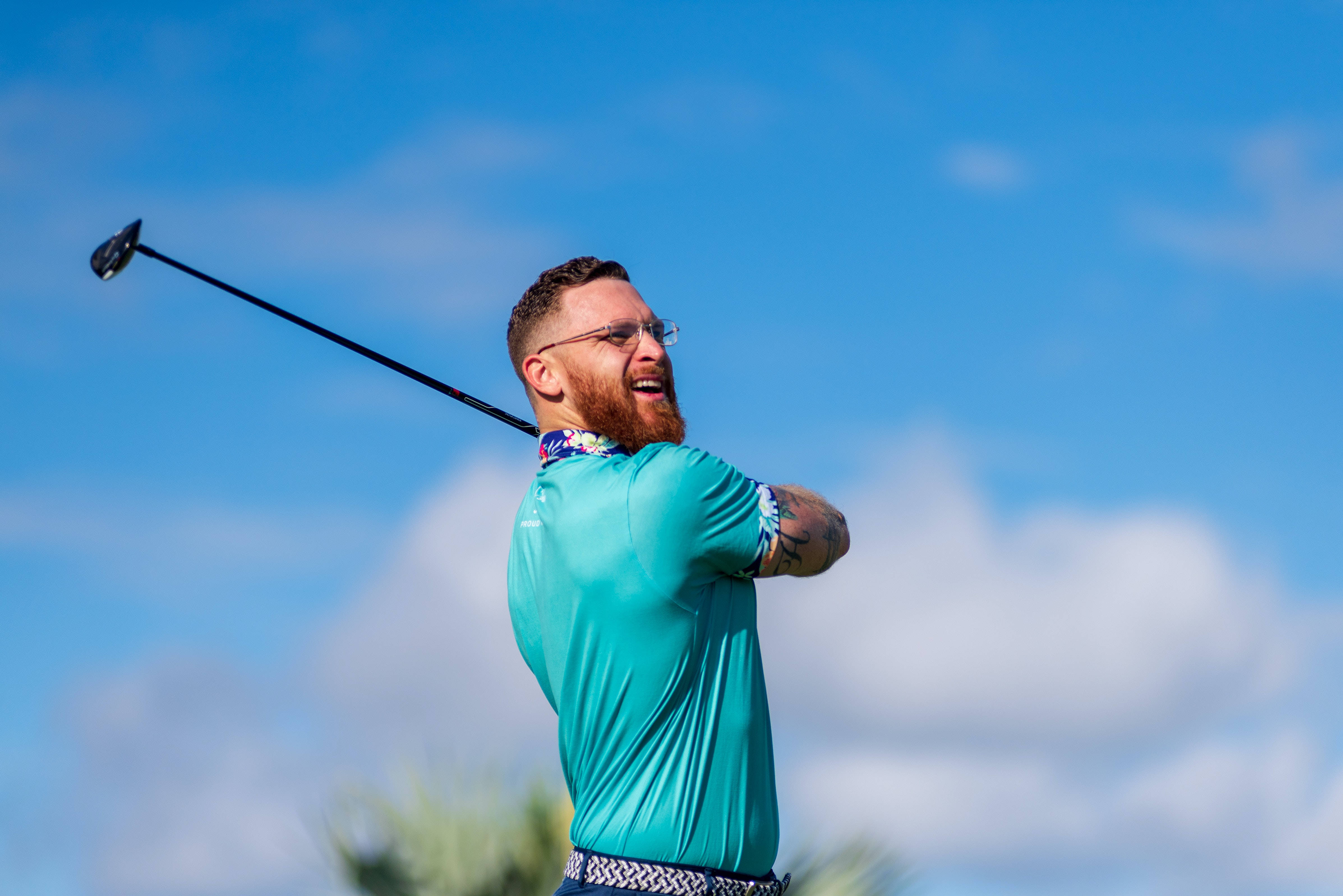 4K Golf 桌布 壁紙 — 一位留鬍鬚戴眼鏡的男子，在藍天白雲下揮桿打高爾夫球時開懷大笑。
