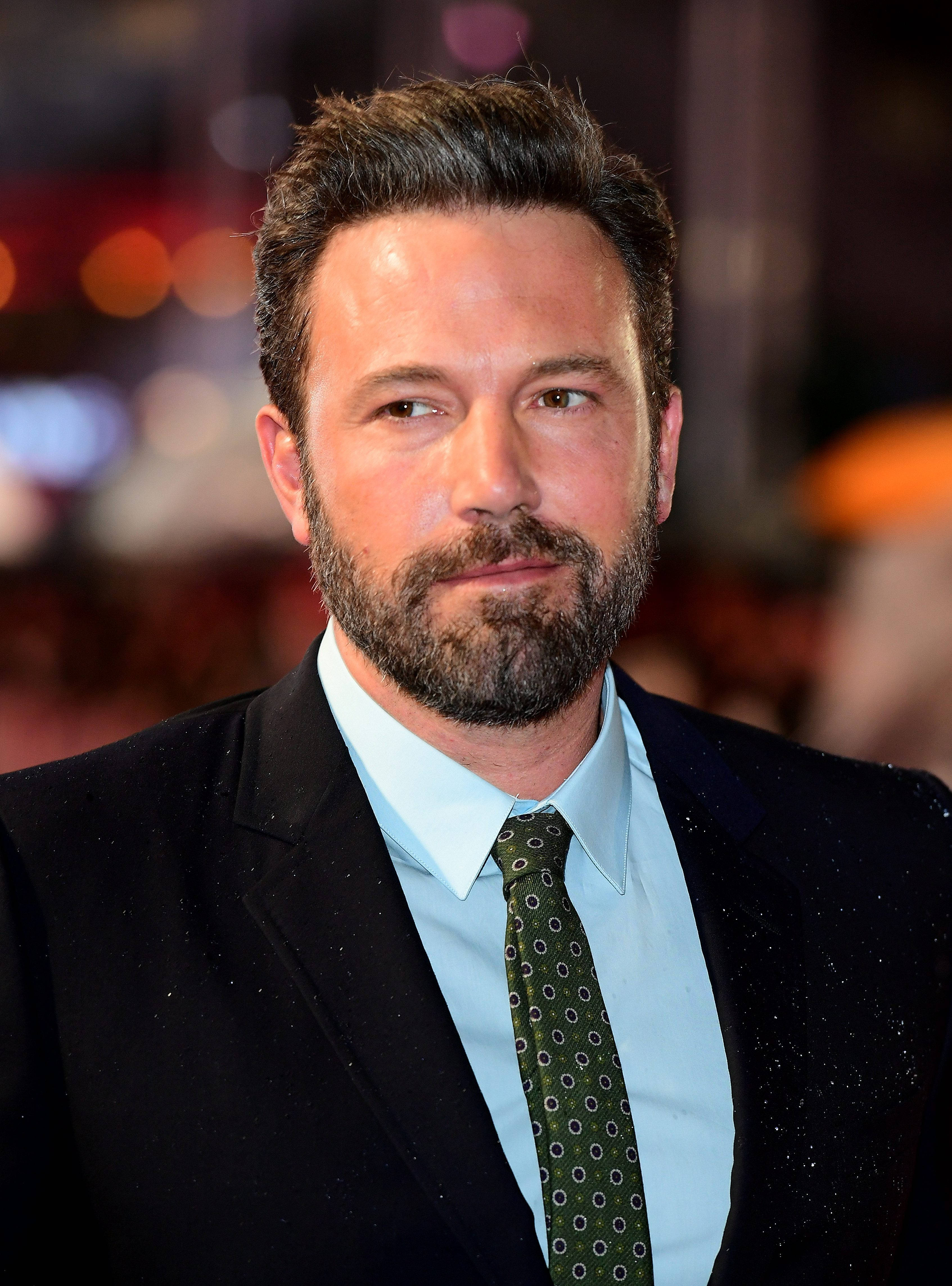 Ben Affleck 桌布 壁紙 — 一位留鬍鬚男士的近距離肖像，身穿黑色西裝和圖案領帶，背景是模糊的城市夜景。