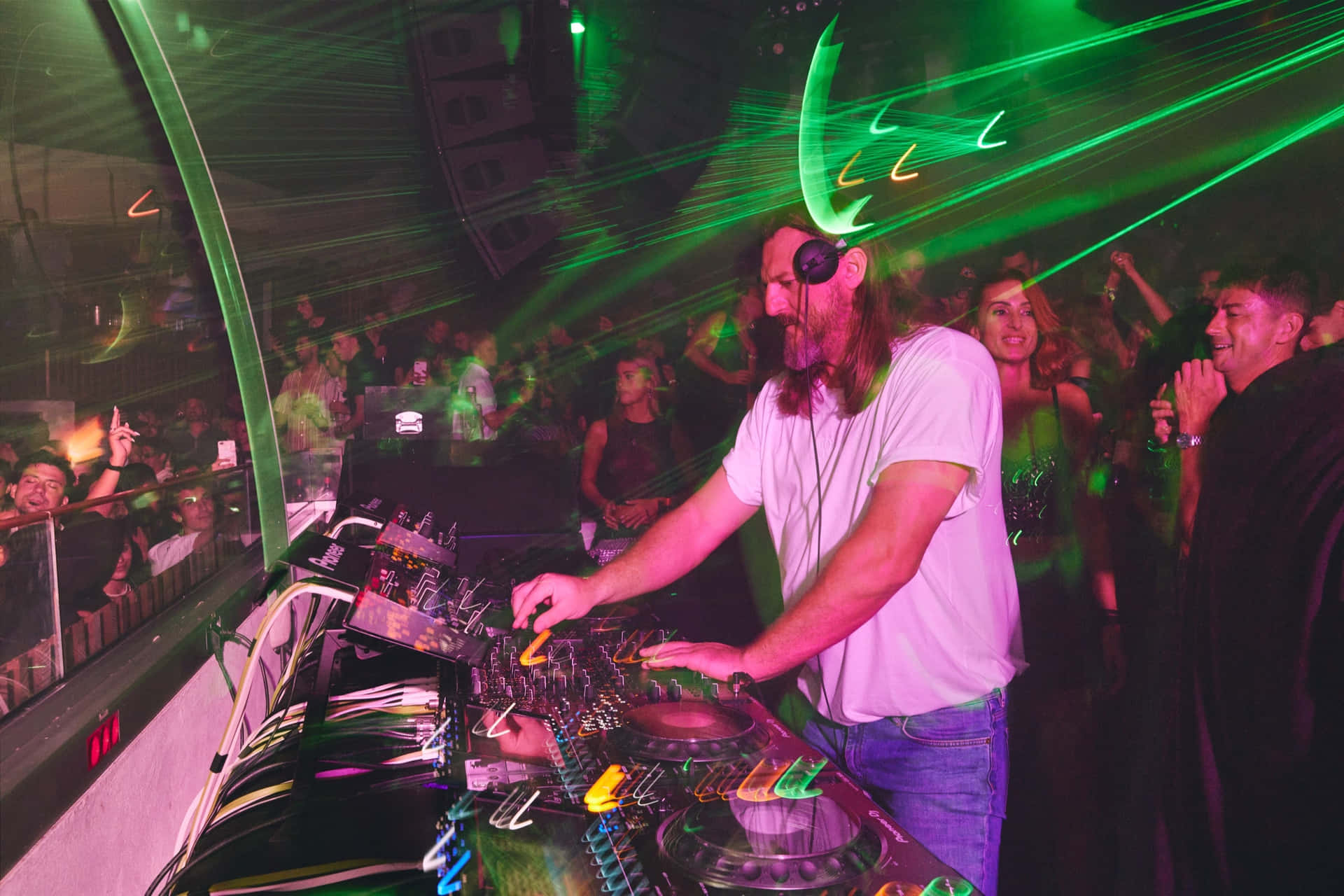 DJ 桌布 壁紙 — 一位留鬍鬚的DJ穿著白色T恤，在充滿活力的綠色雷射燈光下混音，人群在擁擠的夜店中跳舞。