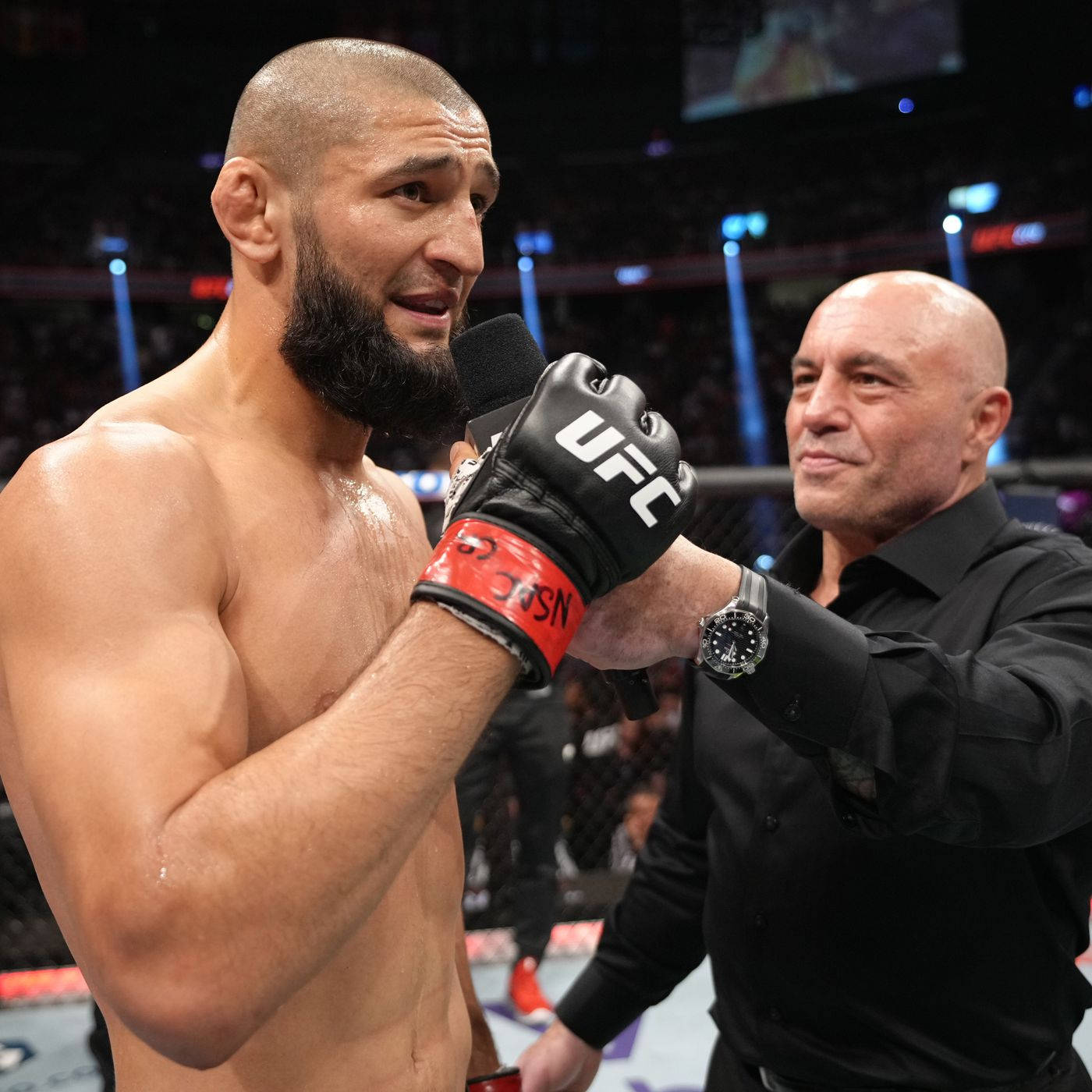 UFC 桌布 壁紙 — 一位留鬍鬚的勝利UFC拳手在八角籠中拿著麥克風，一名穿黑襯衫的光頭男子正在採訪他。