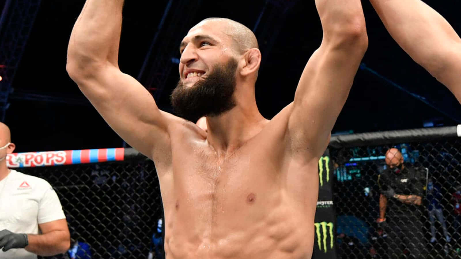 UFC 桌布 壁紙 — 一位留鬍鬚的勝利混合武術家在燈光明亮的八角籠內高舉雙臂慶祝勝利。
