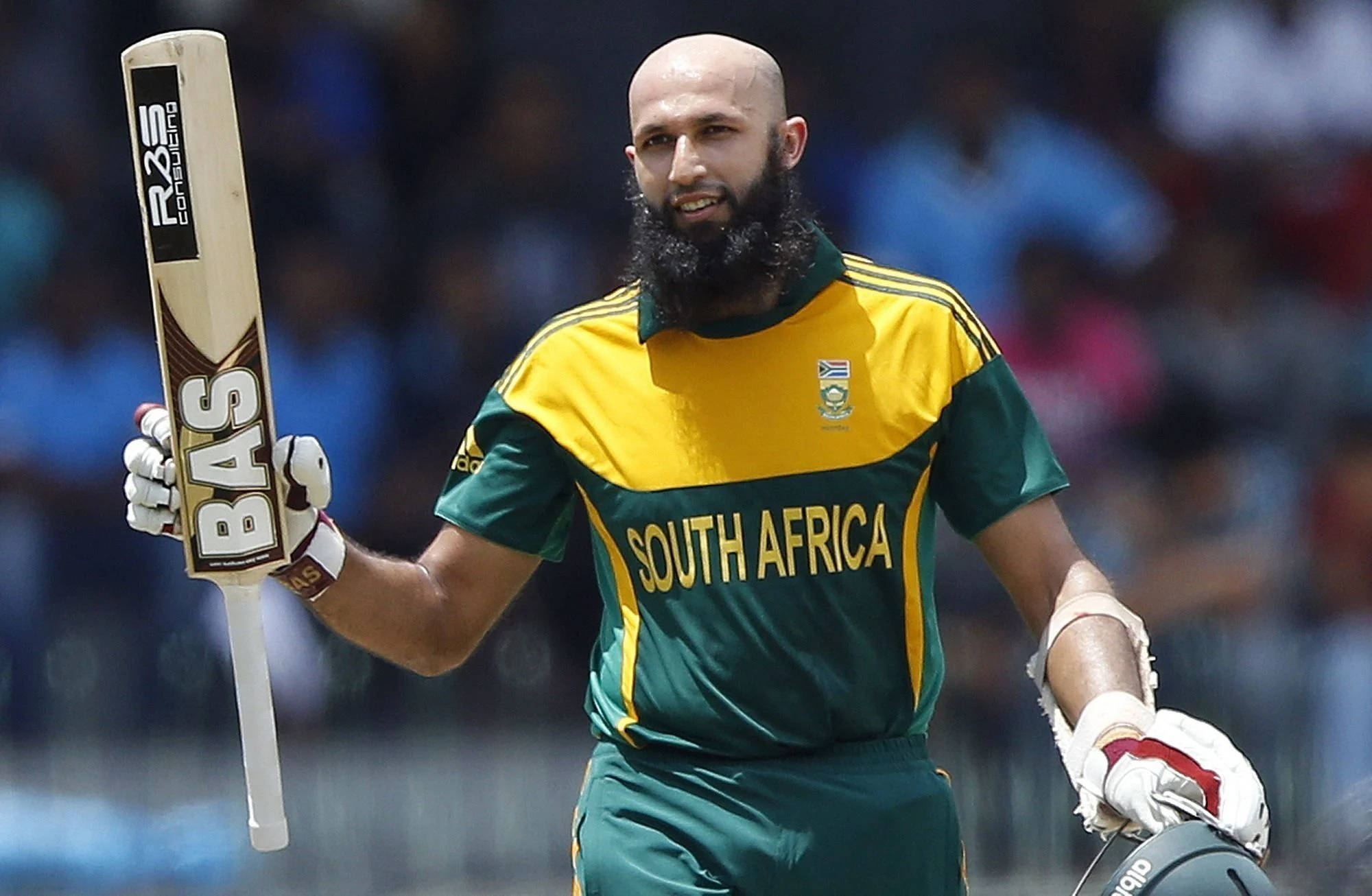 south africa cricket 桌布 壁紙 — 一位留鬍鬚的南非板球運動員在陽光明媚的球場上行走，手持球棒和頭盔。