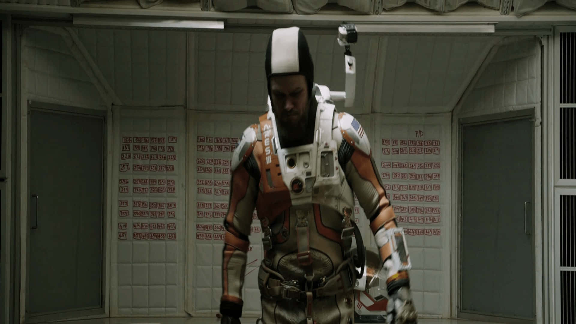 The Martian 桌布 壁紙 — 一位留鬍鬚的太空人穿著細節豐富的橙色和白色太空衣，走過一條牆上有紅色文字的無菌白色走廊。
