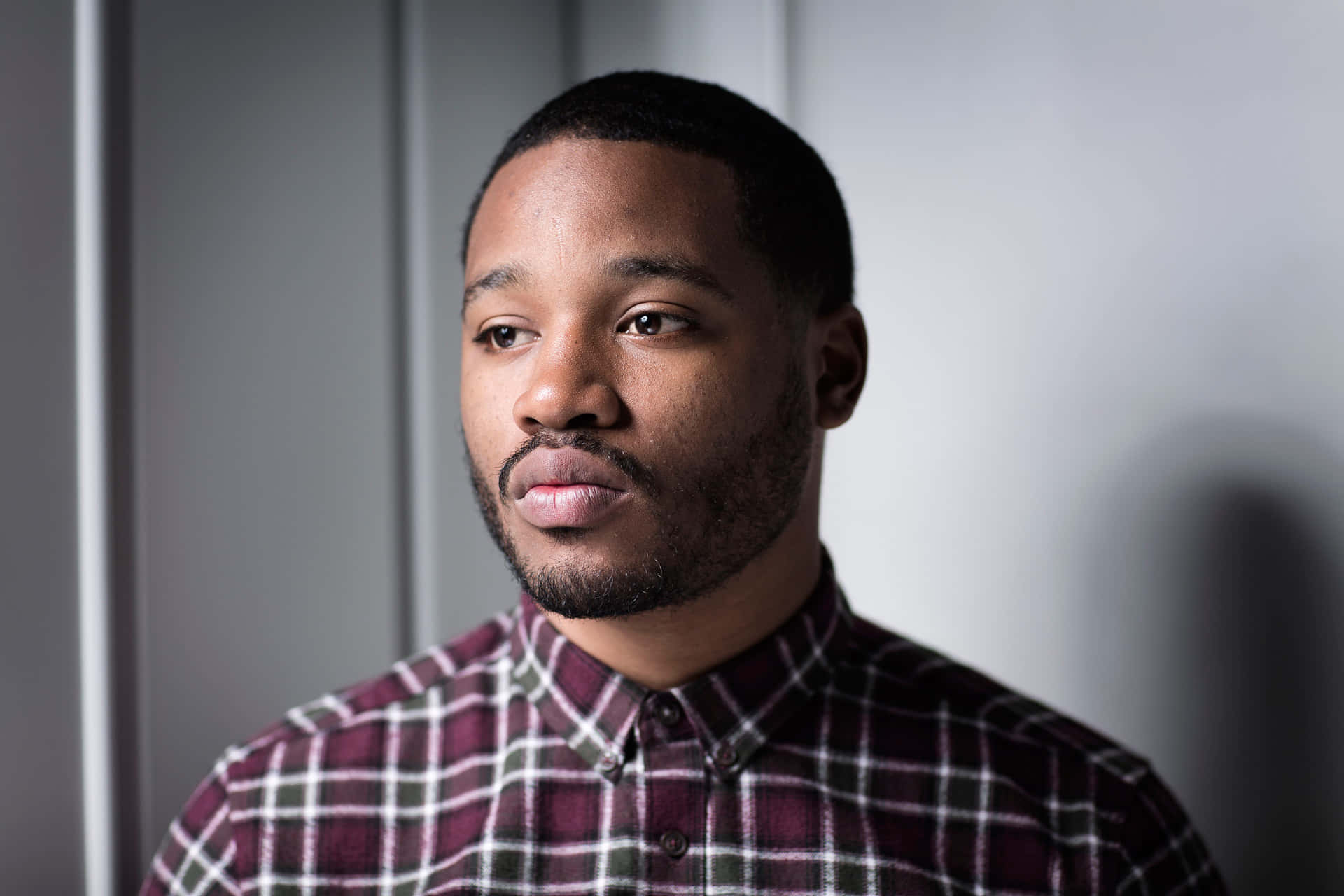 Ryan Coogler 桌布 壁紙 — 一位留鬍鬚的年輕男子穿著栗紅色格紋襯衫，背景是簡約的灰色。