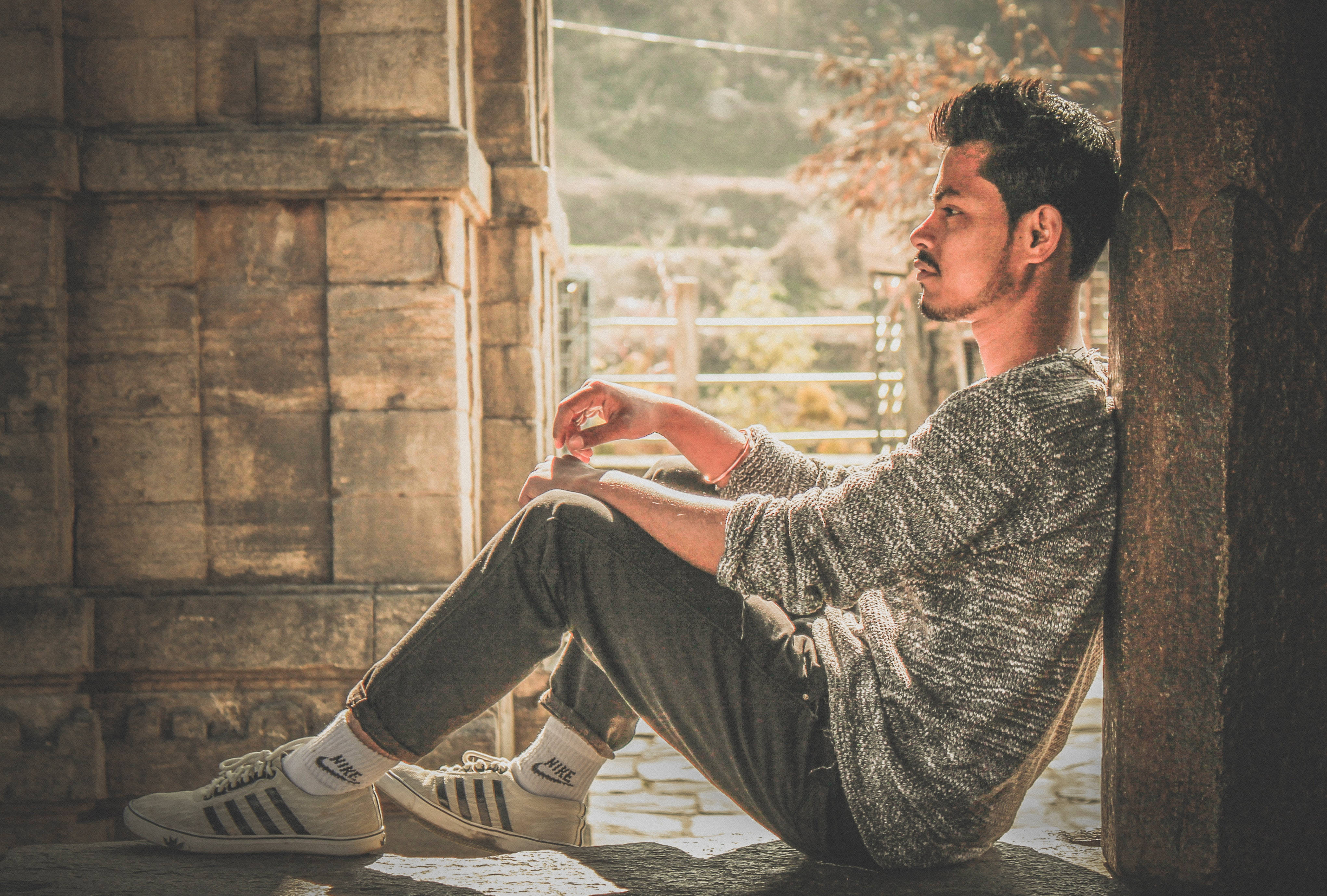 male model 桌布 壁紙 — 一位留鬍鬚的年輕男子靠著石柱沉思，石柱沐浴在穿過樹葉的溫暖金色陽光中。