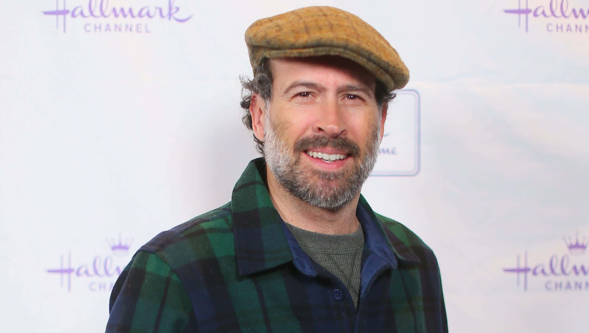 Jason Lee 桌布 壁紙 — 一位留鬍鬚的微笑男士戴著棕色平頂帽和綠色格紋襯衫，背景是 Hallmark Channel。