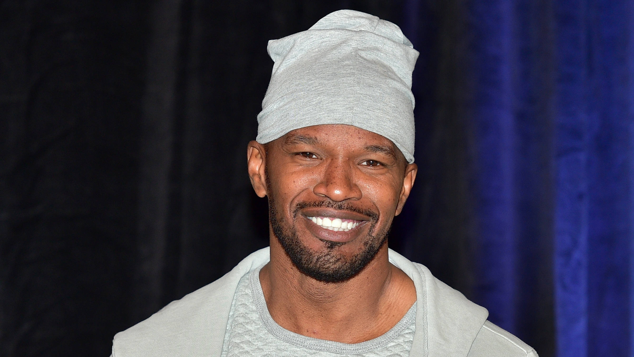Jamie Foxx 桌布 壁紙 — 一位留鬍鬚的微笑男士戴著灰色毛帽和連帽上衣，背景是深藍色窗簾。