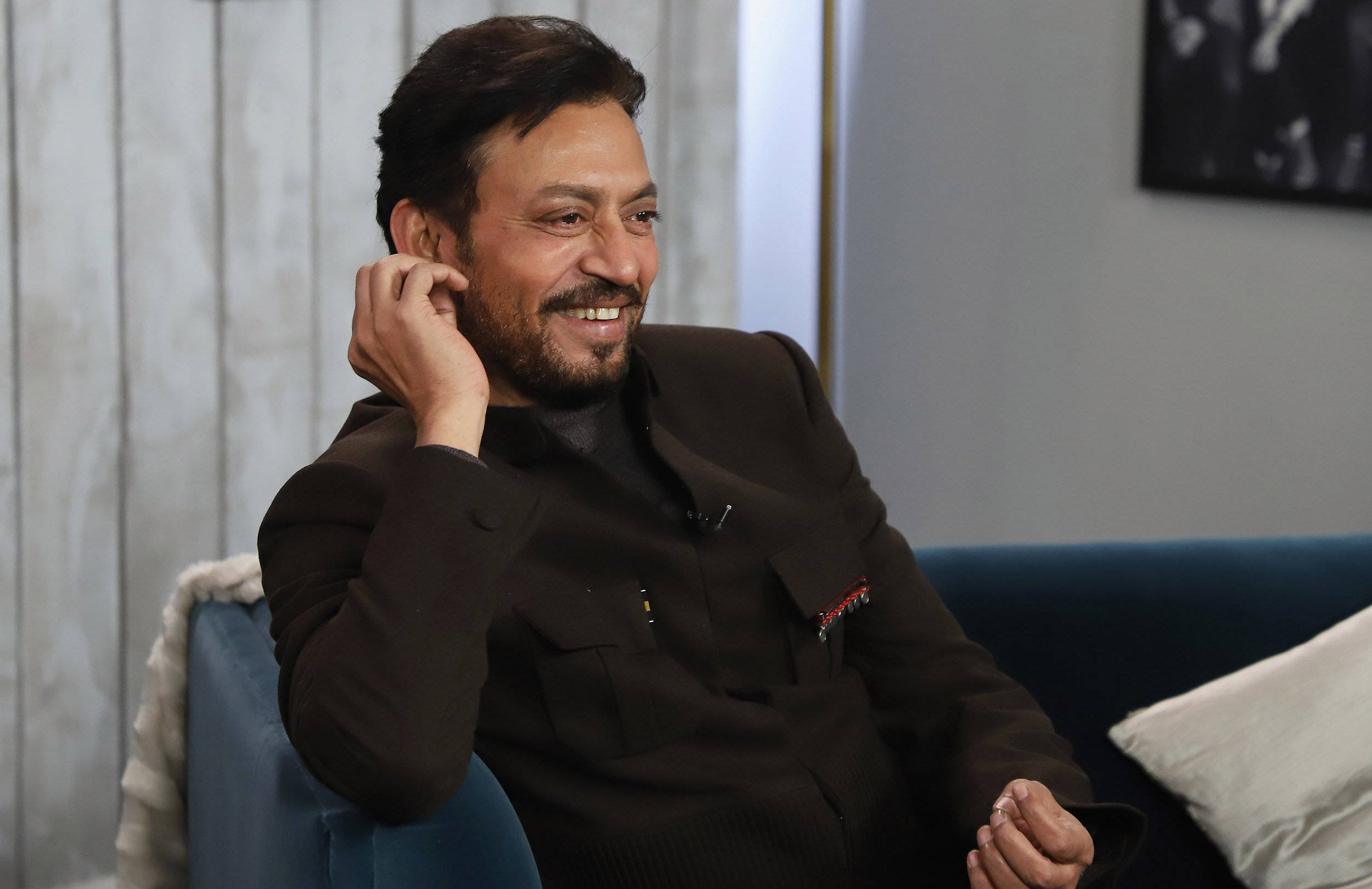 Irrfan Khan 桌布 壁紙 — 一位留鬍鬚的微笑男士放鬆地坐在現代室內設計的藍色沙發上，手靠近耳朵。