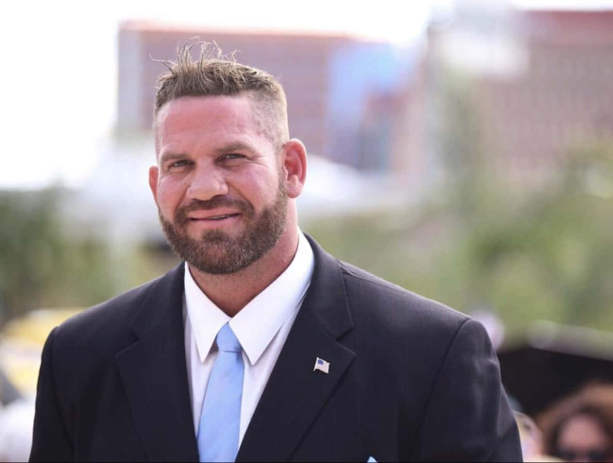 WWE 桌布 壁紙 — 一位留鬍鬚的微笑男士穿著深色西裝和淺藍色領帶，背景是模糊的城市景觀。