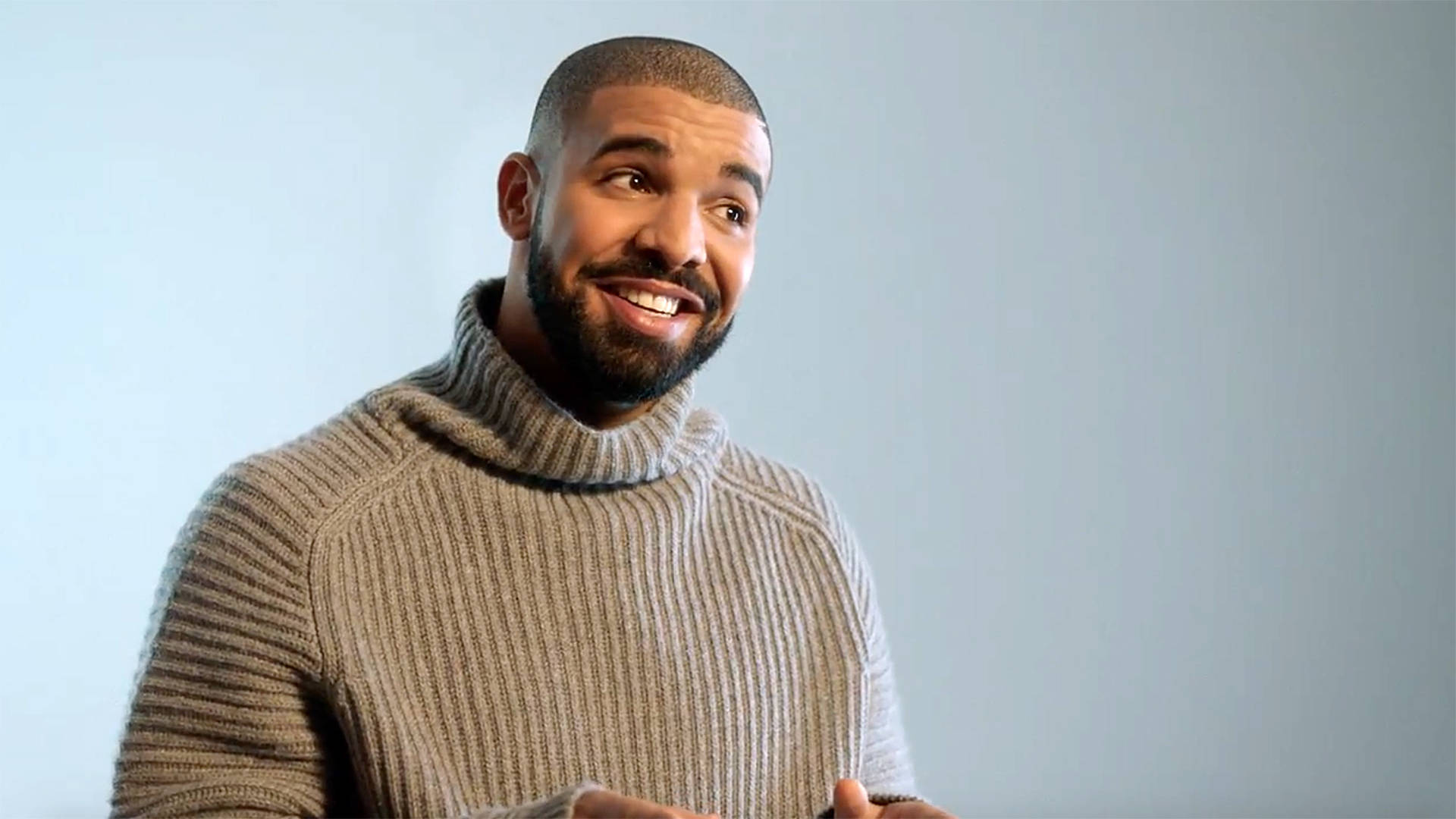 Drake 桌布 壁紙 — 一位留鬍鬚的微笑男士穿著羅紋高領毛衣，背景是純淺藍色。