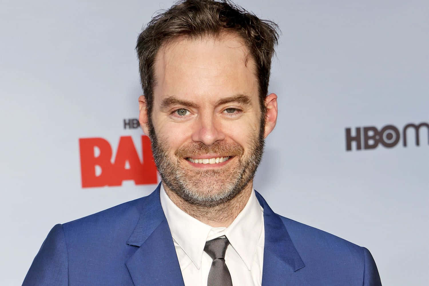 bill hader 桌布 壁紙 — 一位留鬍鬚的微笑男士穿著藍色西裝和灰色領帶，背景是HBO Max。