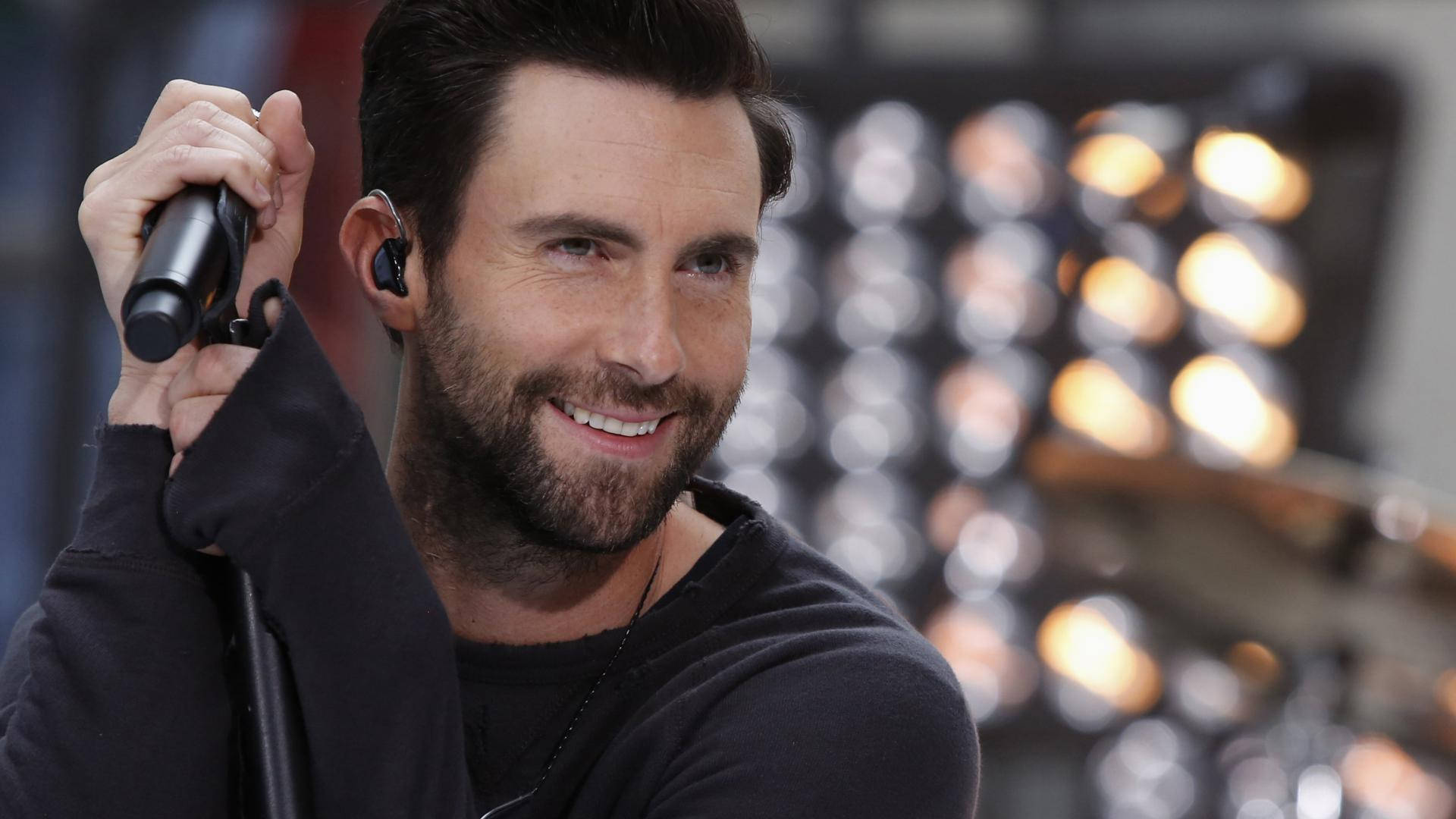 Adam Levine 桌布 壁紙 — 一位留鬍鬚的微笑男子戴著耳機，在閃爍的舞台燈光背景下握著麥克風。