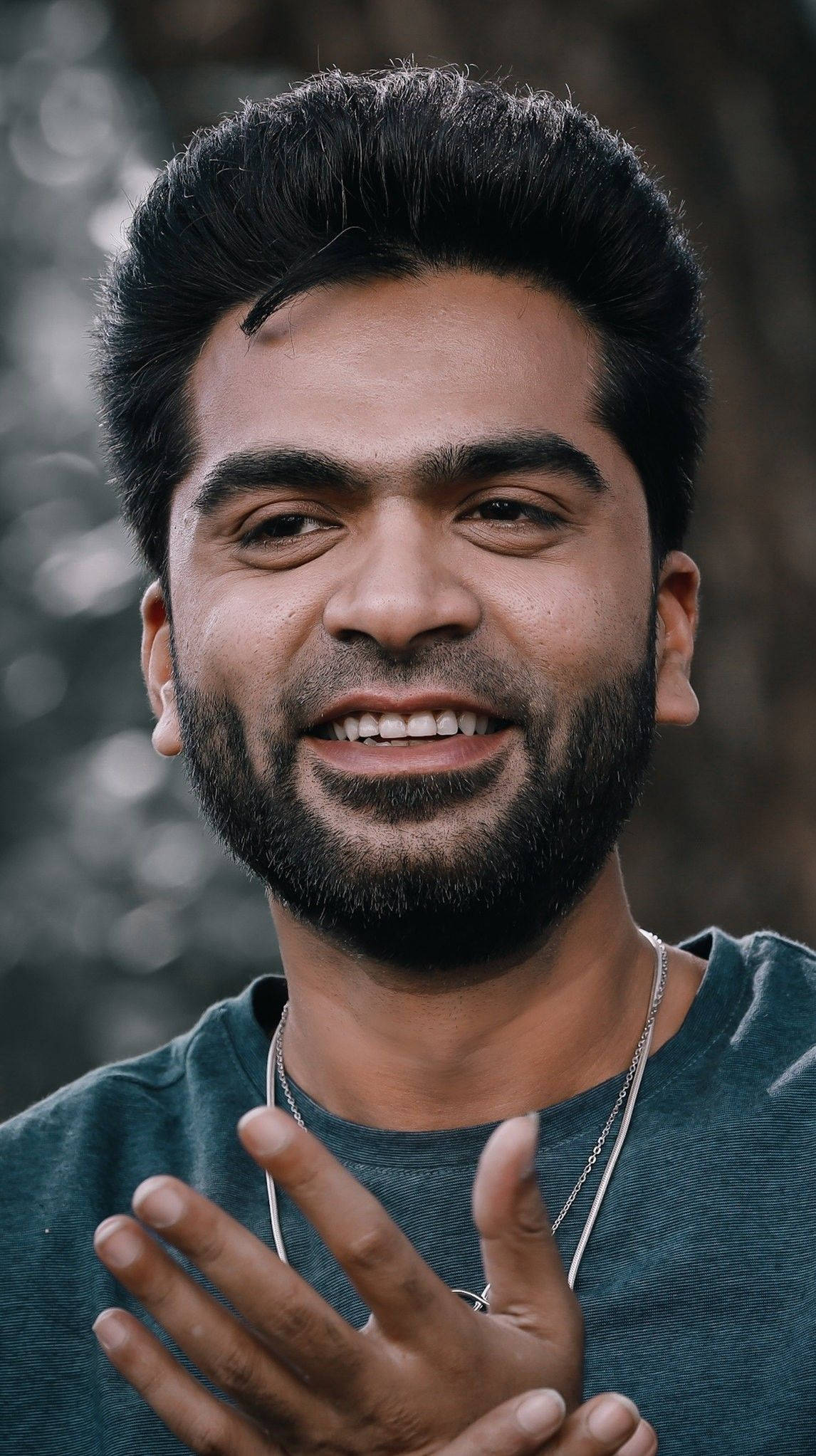 Indian 桌布 壁紙 — 一位留鬍鬚的快樂男子，在模糊的自然背景下溫暖微笑並用手勢表達。