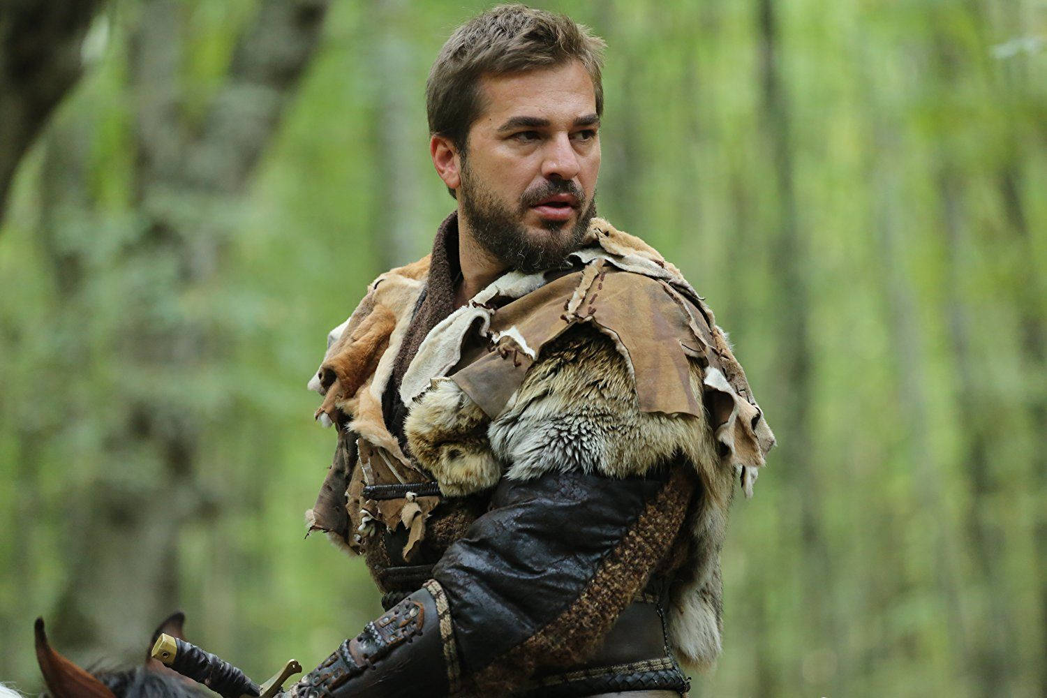 Ertugrul 桌布 壁紙 — 一位留鬍鬚的戰士穿著毛皮襯裡的盔甲，騎著馬穿過茂密的綠色森林。