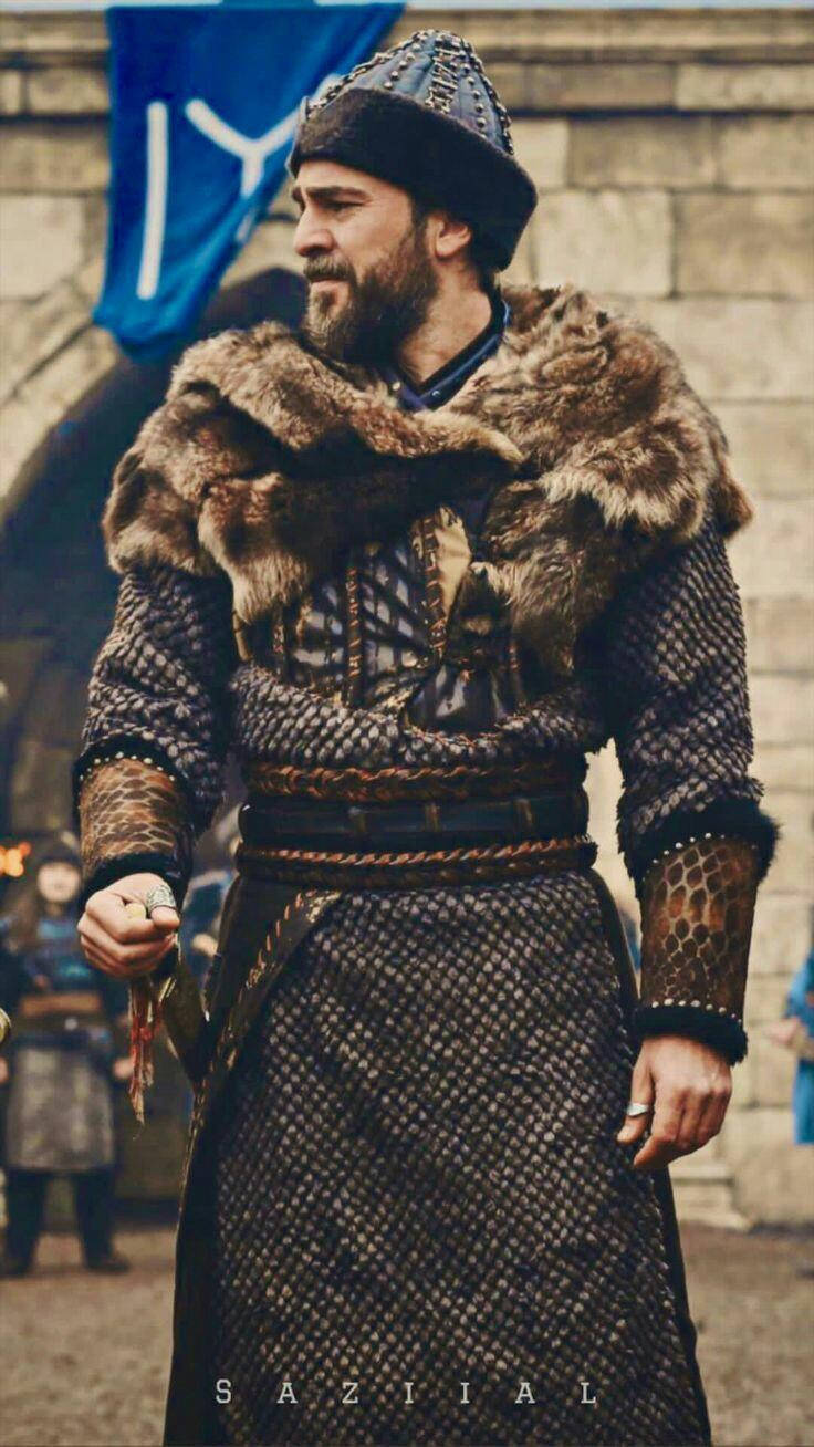 Ertugrul 桌布 壁紙 — 一位留鬍鬚的戰士穿著毛皮襯裡的鎖子甲，站在藍色旗幟下的石城堡門前。
