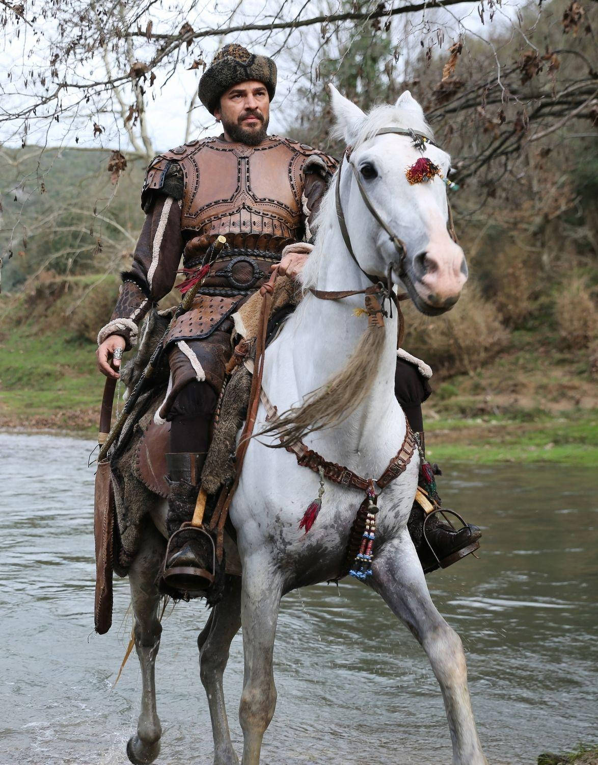 Ertugrul 桌布 壁紙 — 一位留鬍鬚的戰士穿著精細的盔甲，騎著裝飾的白馬，在光禿禿的樹木下穿過淺淺的河水。