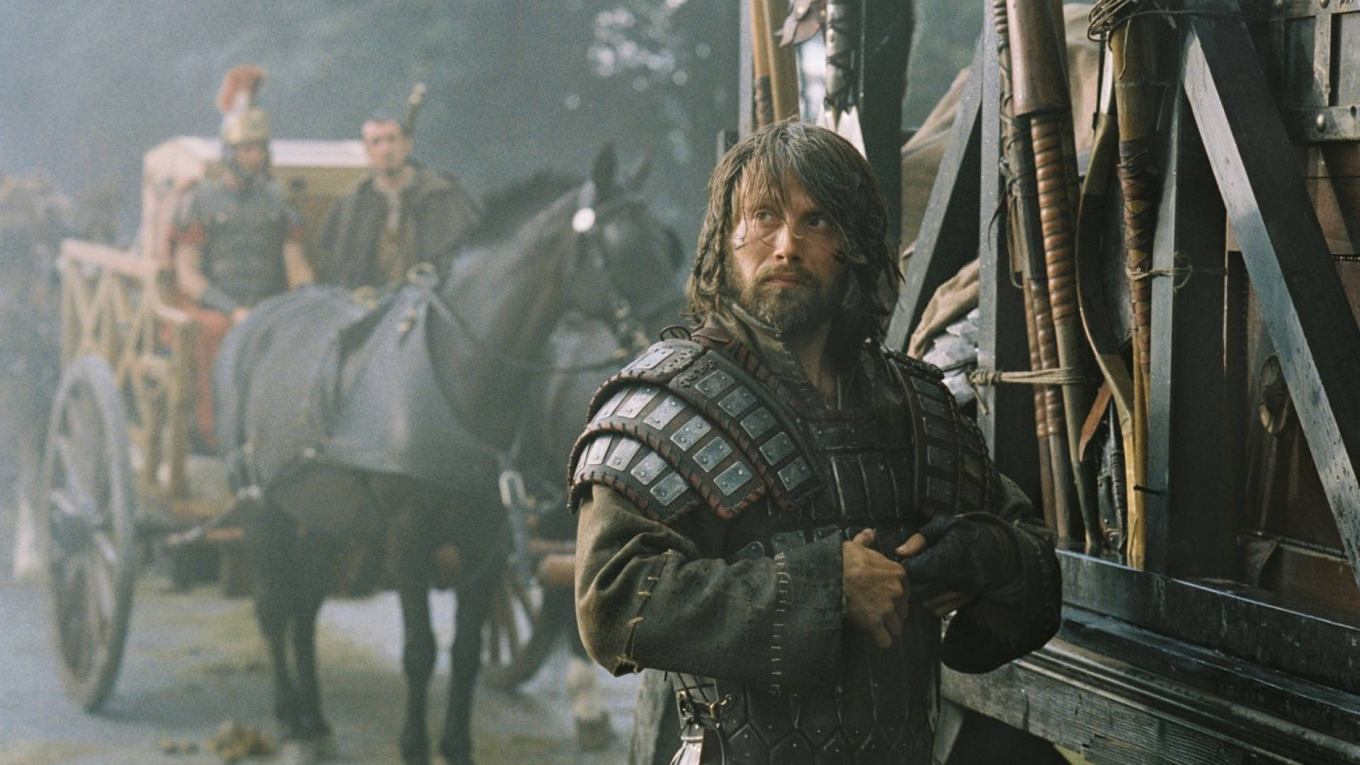 Mads Mikkelsen 桌布 壁紙 — 一位留鬍鬚的戰士身穿分段式鎧甲，堅定地站在一輛由黑馬拉動的木製戰車旁，身處迷霧戰場。
