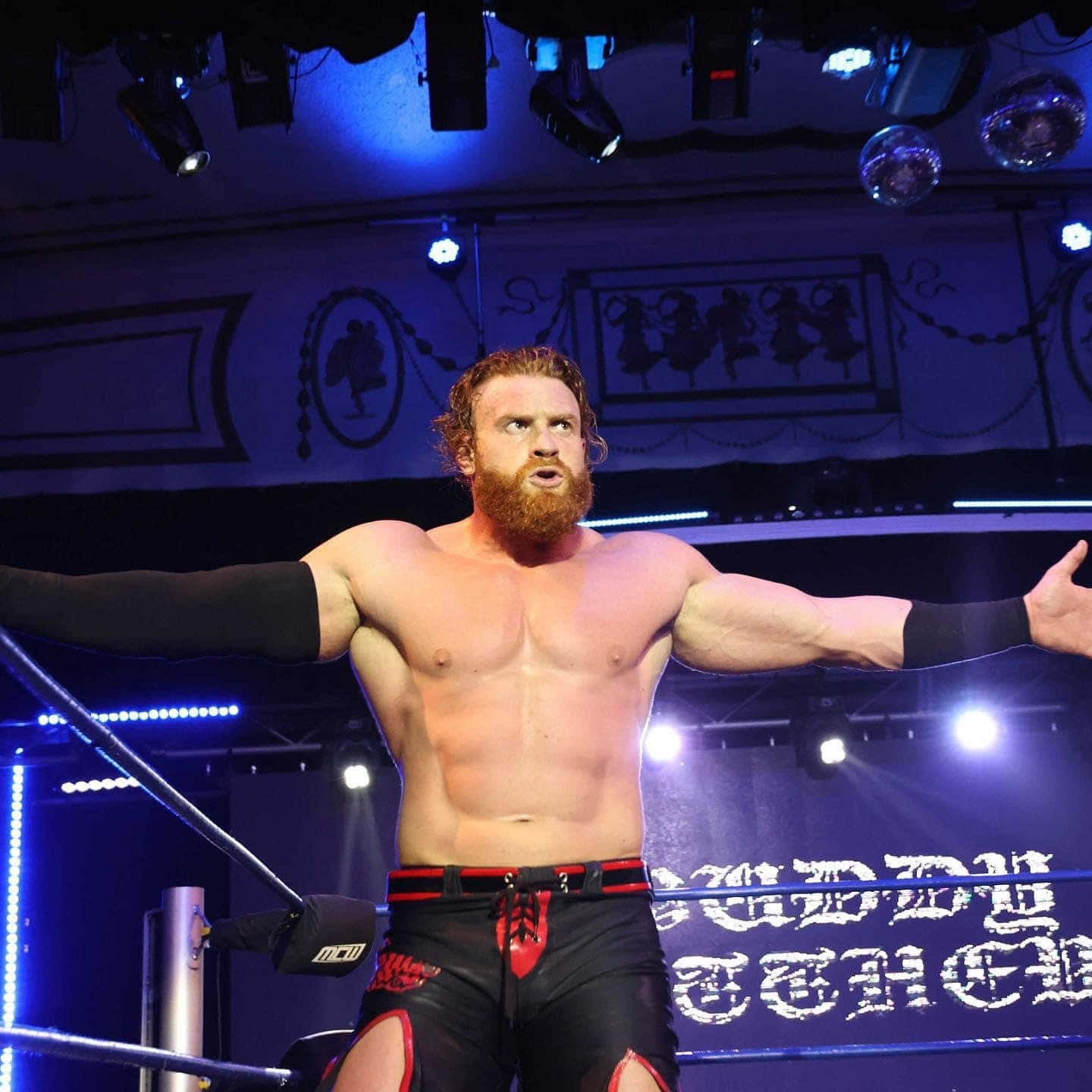 WWE 桌布 壁紙 — 一位留鬍鬚的摔角手穿著黑色和紅色的短褲，在藍色舞台燈光下張開雙臂，勝利地站在擂台上。