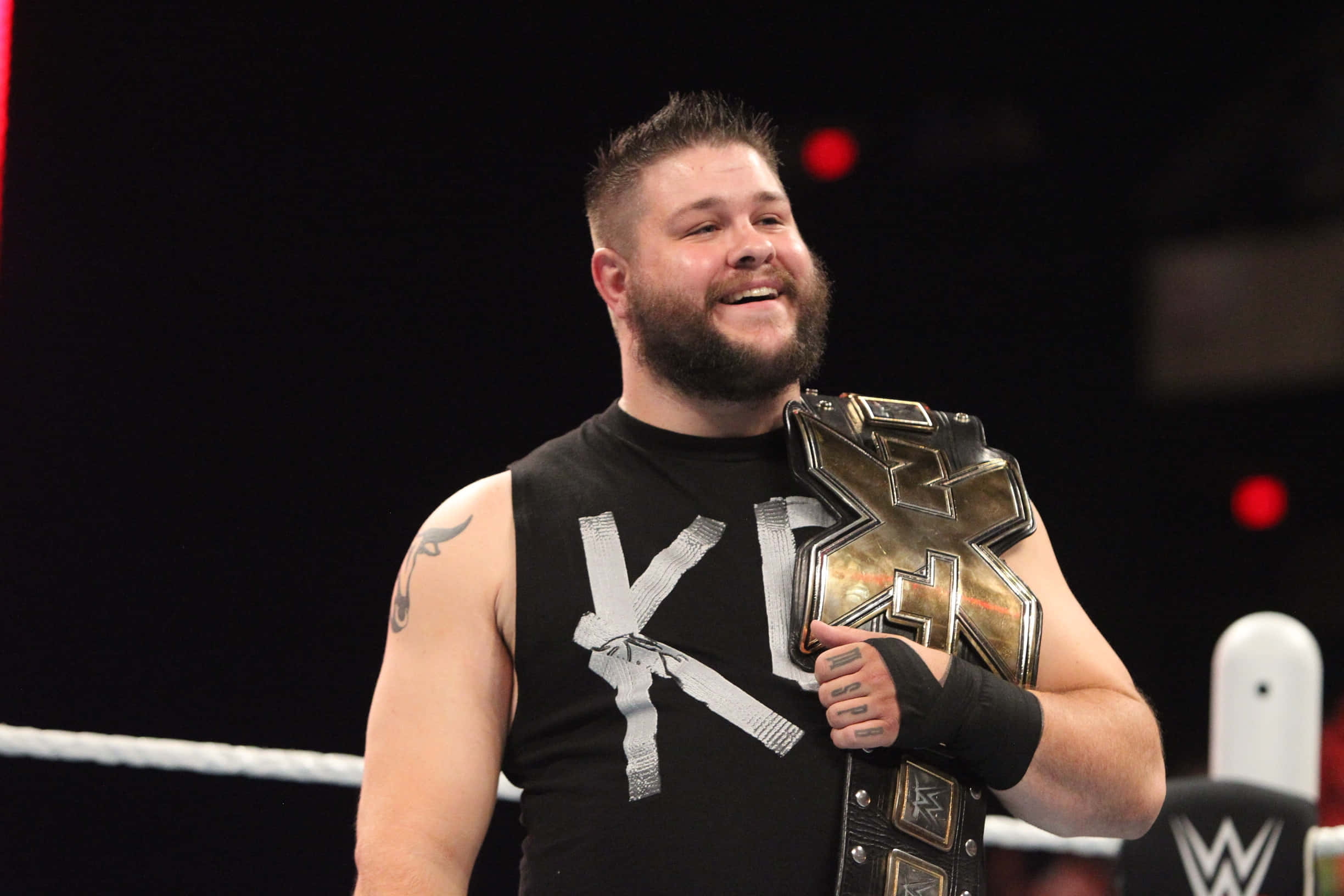 WWE 桌布 壁紙 — 一位留鬍鬚的摔角手身穿黑色背心，在摔角場內手持冠軍腰帶，笑容滿面。