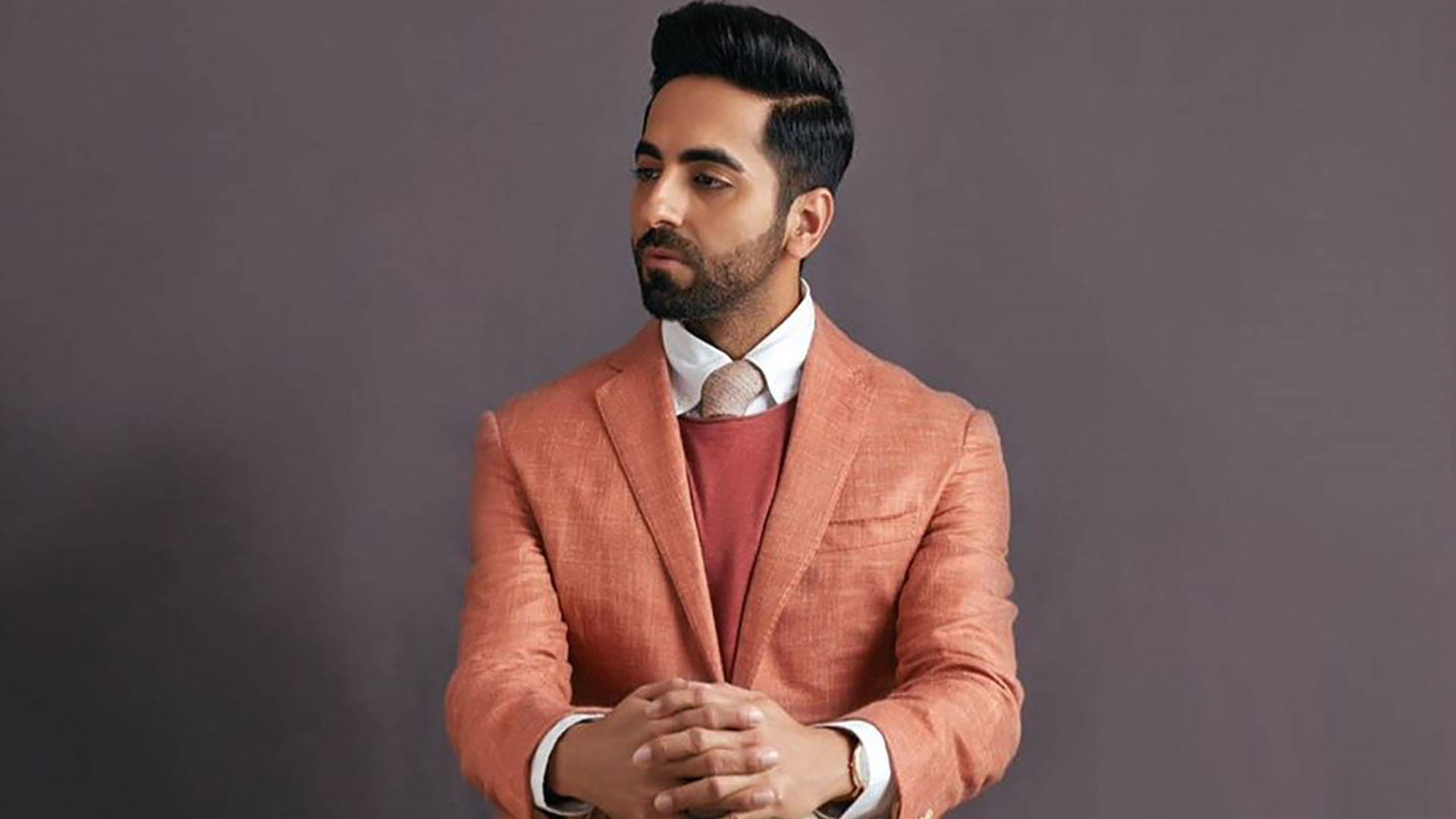 ayushmann khurrana 桌布 壁紙 — 一位留鬍鬚的時尚男士，在柔和的灰色工作室背景前，穿著珊瑚色西裝外套，內搭栗色毛衣。