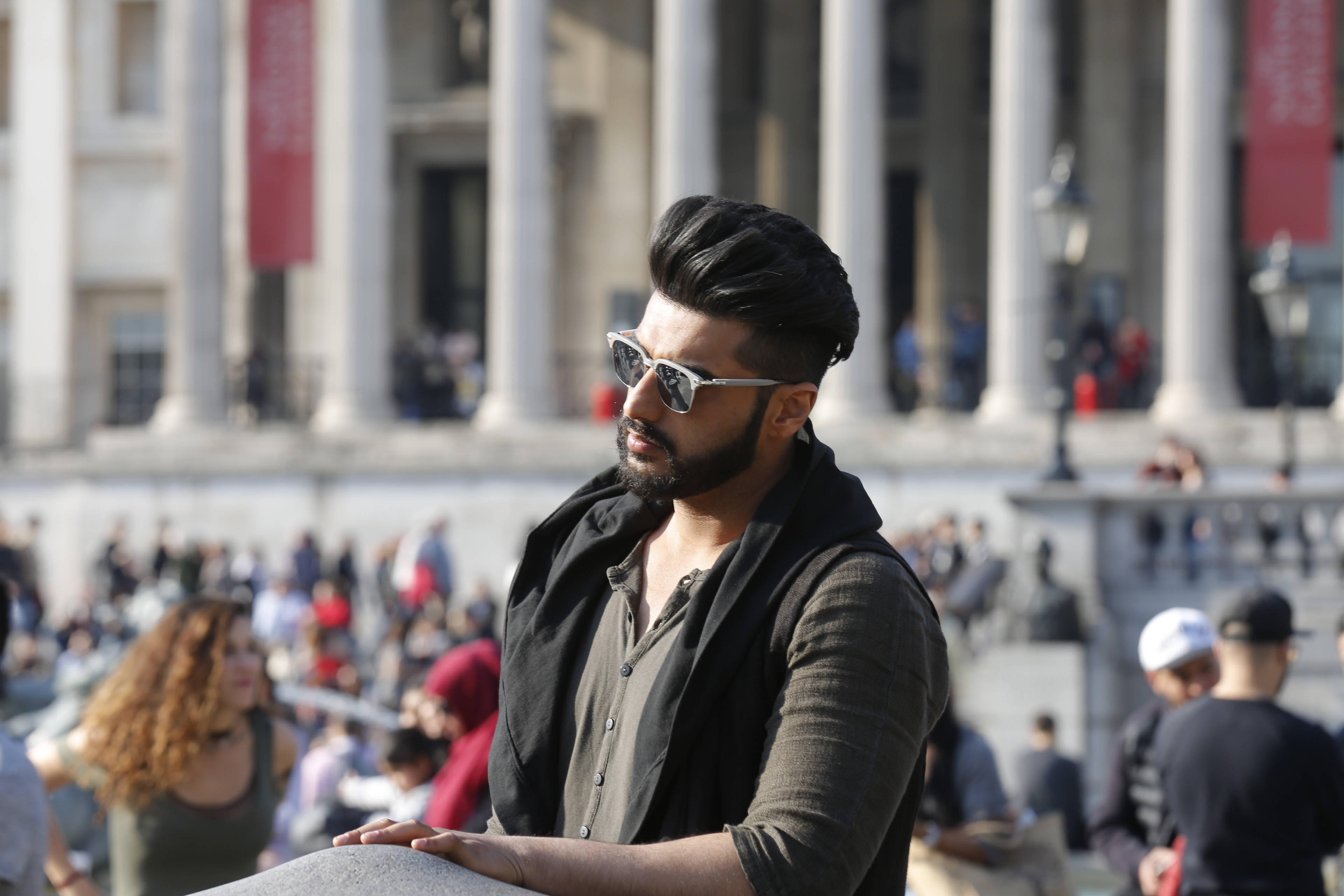 Arjun Kapoor 桌布 壁紙 — 一位留鬍鬚的時尚男士戴著太陽眼鏡，靠在高柱宏偉古典建築前的石欄杆上。
