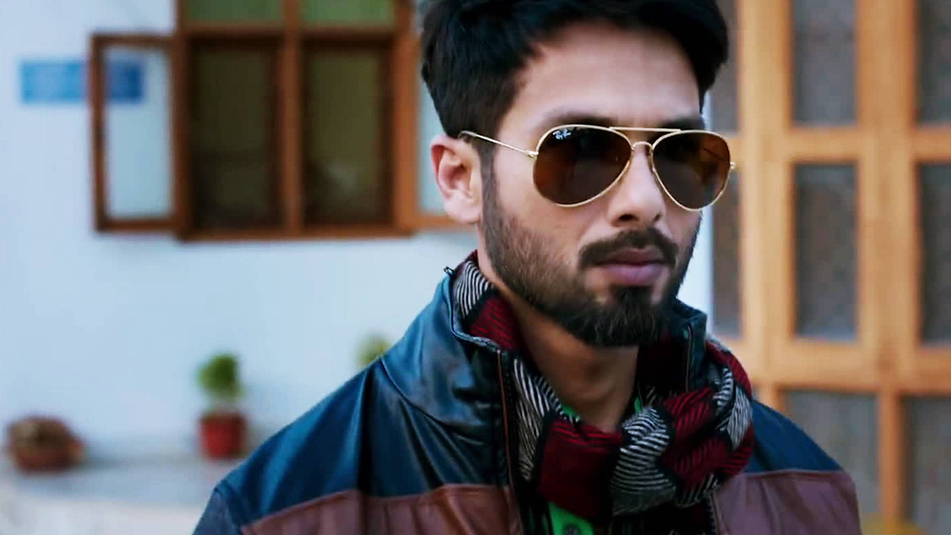 Shahid Kapoor 桌布 壁紙 — 一位留鬍鬚的時尚男士戴著金色邊框的飛行員太陽眼鏡和彩色圍巾，以木窗為背景。