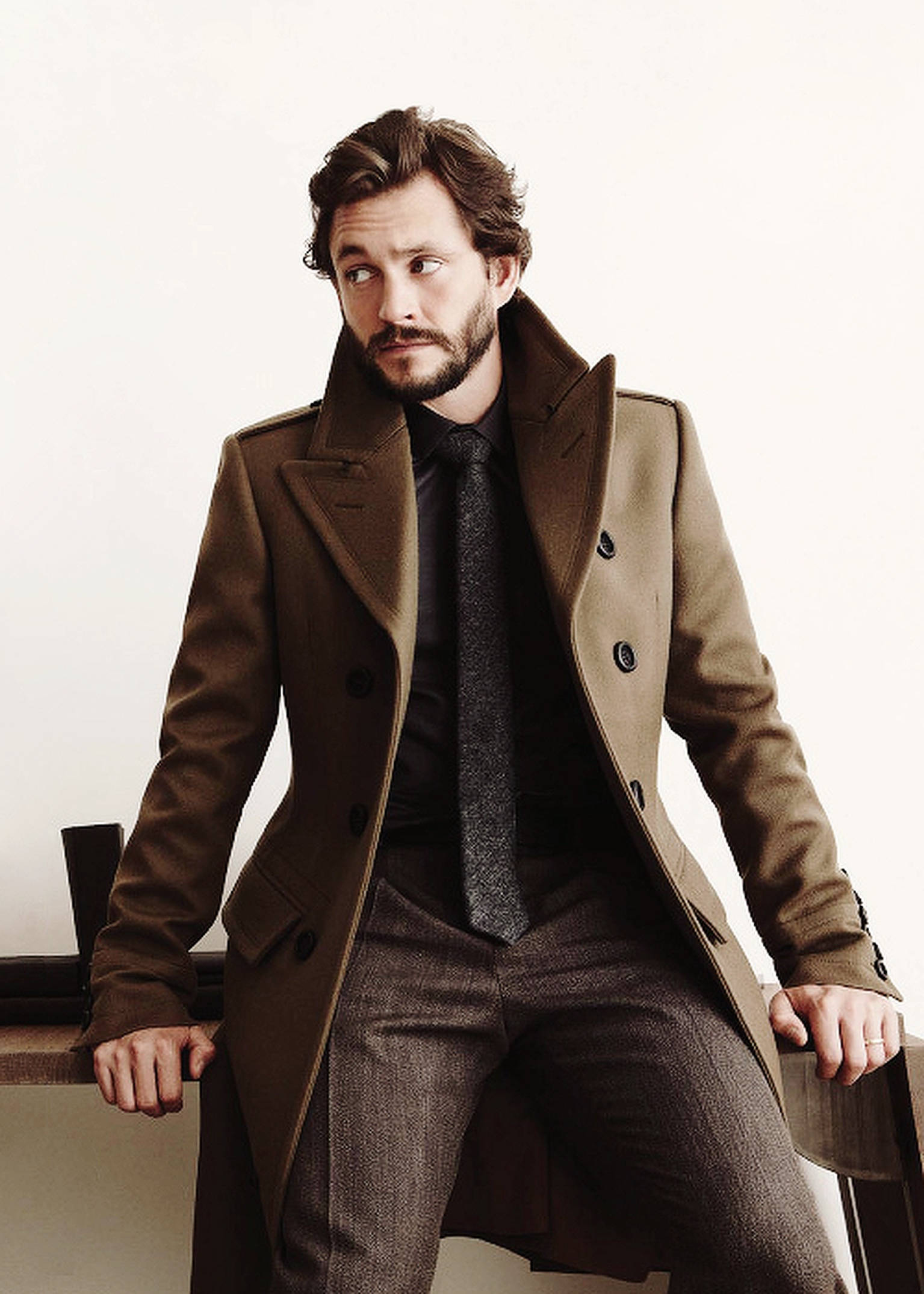 Hugh Dancy 桌布 壁紙 — 一位留鬍鬚的時尚男士穿著棕色外套，自信地坐在白色牆壁前的木長椅上。