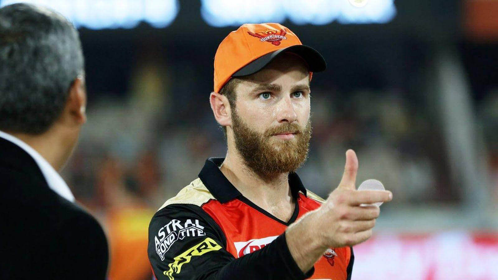 kane williamson 桌布 壁紙 — 一位留鬍鬚的板球運動員，戴著橙色帽子和球衣，在夜間比賽中在體育場燈光下豎起大拇指。