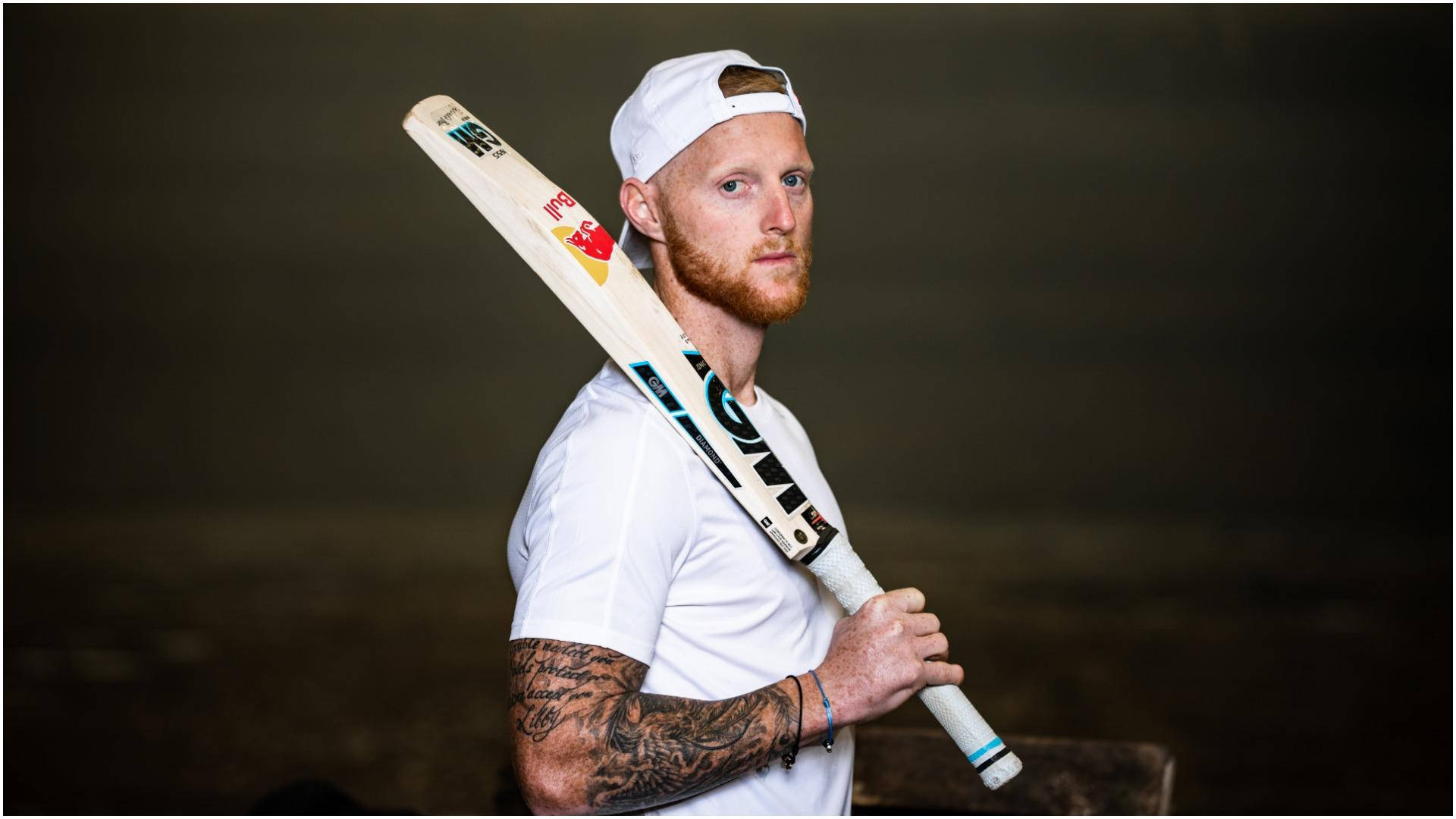 Ben Stokes 桌布 壁紙 — 一位留鬍鬚的板球運動員身穿白色裝備，在黑暗模糊的背景中將球棒扛在肩上。