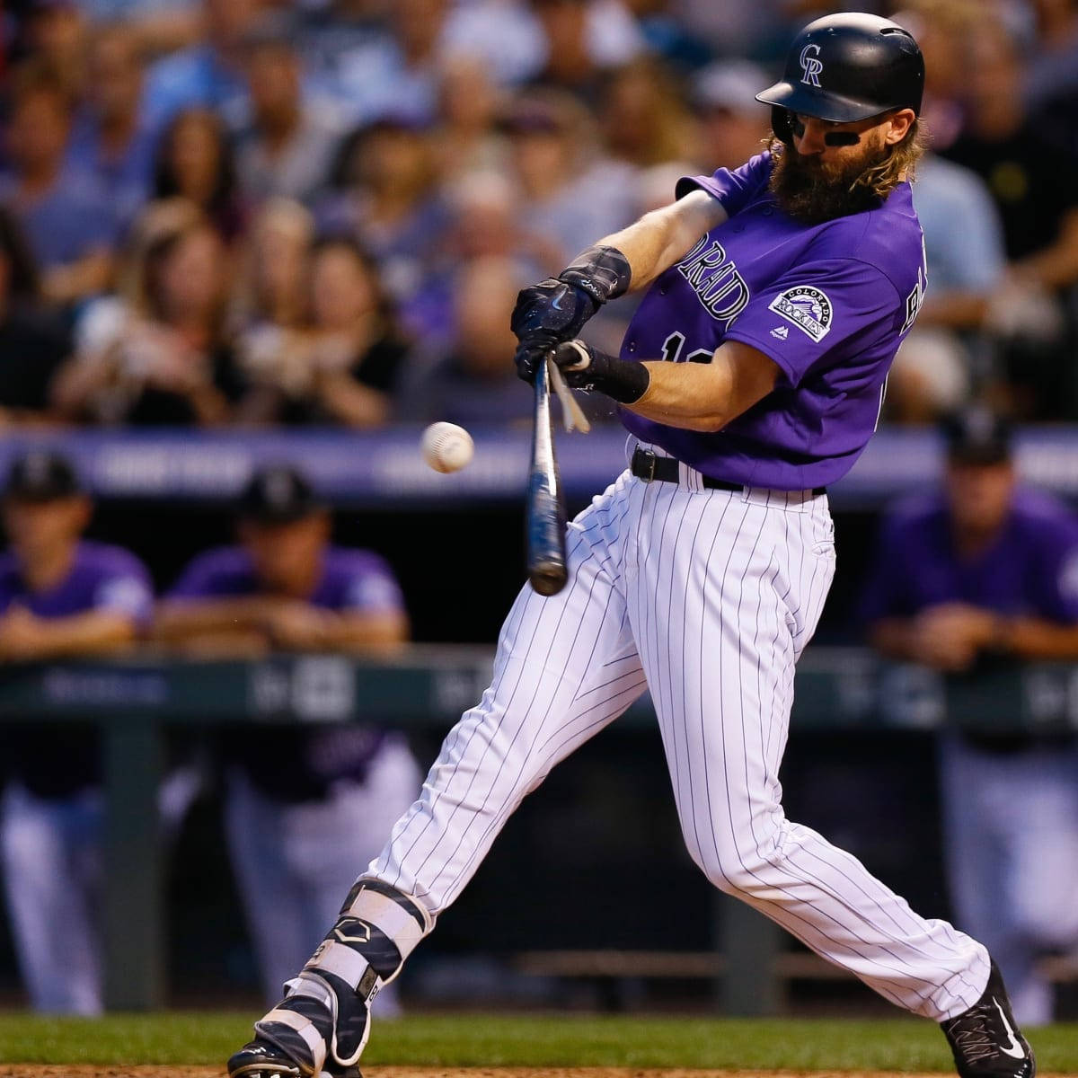運動 桌布 壁紙 — 一位留鬍鬚的棒球運動員穿著紫色制服，在夜間比賽中揮棒擊球，背景有模糊的觀眾。
