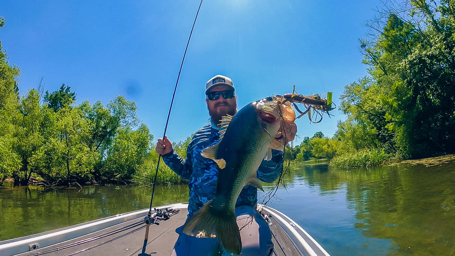 Fishing Rod 桌布 壁紙 — 一位留鬍鬚的漁夫在船上自豪地舉起一條在陽光照耀的河流中捕獲的巨大鱸魚，周圍環繞著茂密的綠樹。
