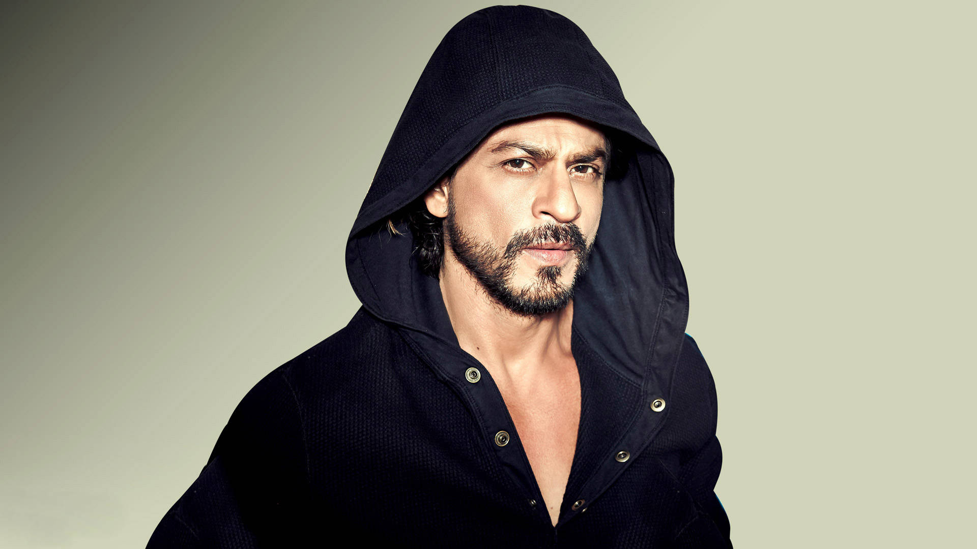 Shahrukh 桌布 壁紙 — 一位留鬍鬚的男人，穿著深色連帽衫，在純色淺色背景前，眼神凌厲。