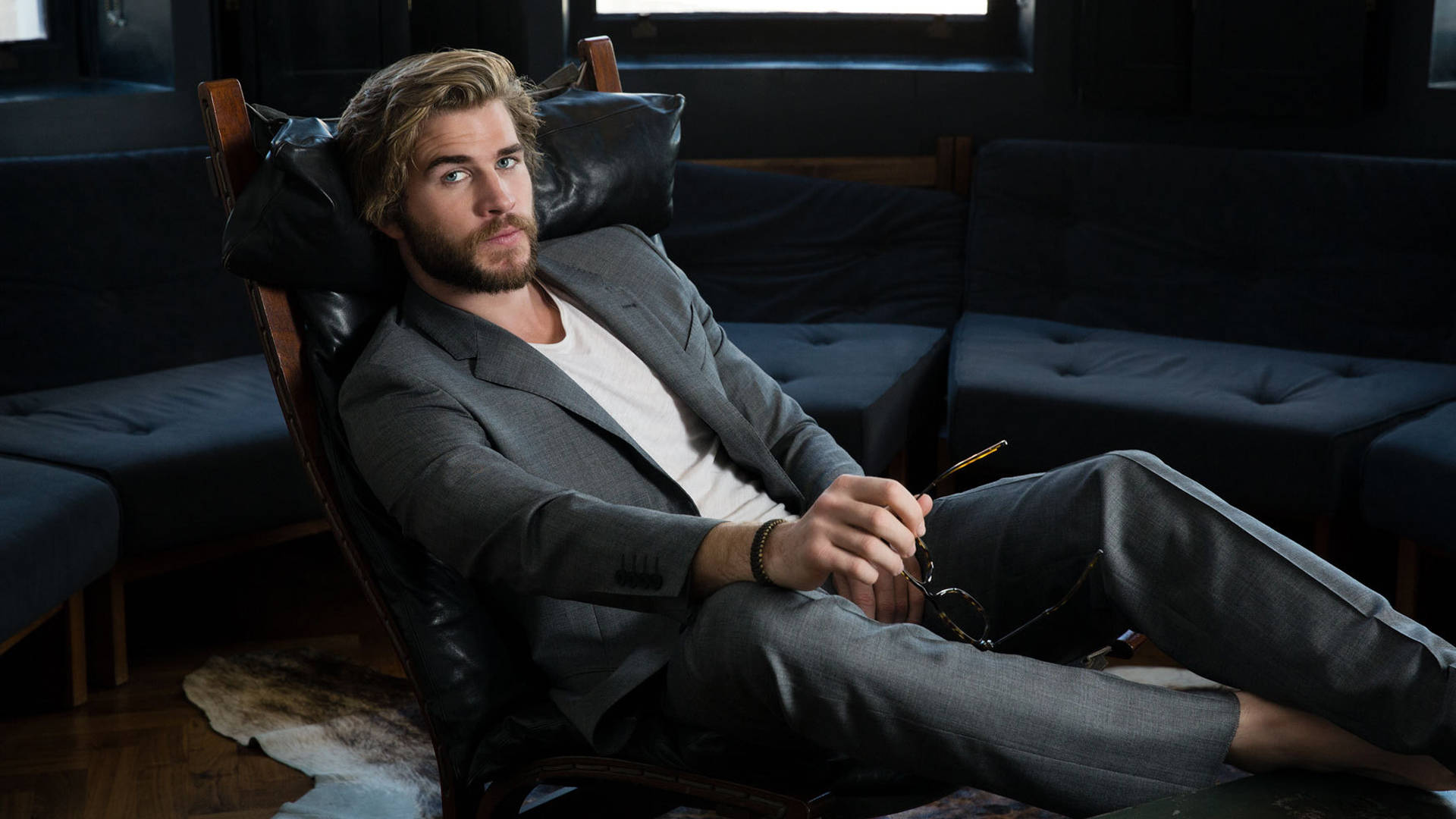 liam 桌布 壁紙 — 一位留鬍鬚的男人在昏暗的房間裡穿著灰色西裝，放鬆地坐在皮革椅上，手持眼鏡。