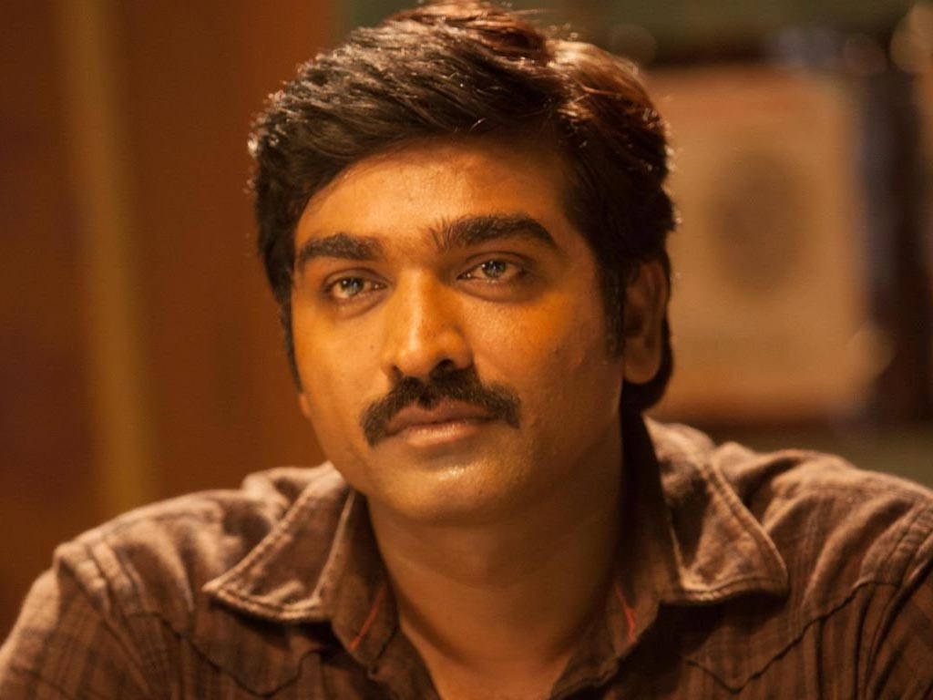 vijay HD 桌布 壁紙 — 一位留鬍鬚的男人在溫暖光線的室內環境中若有所思地凝視，背景柔和模糊。