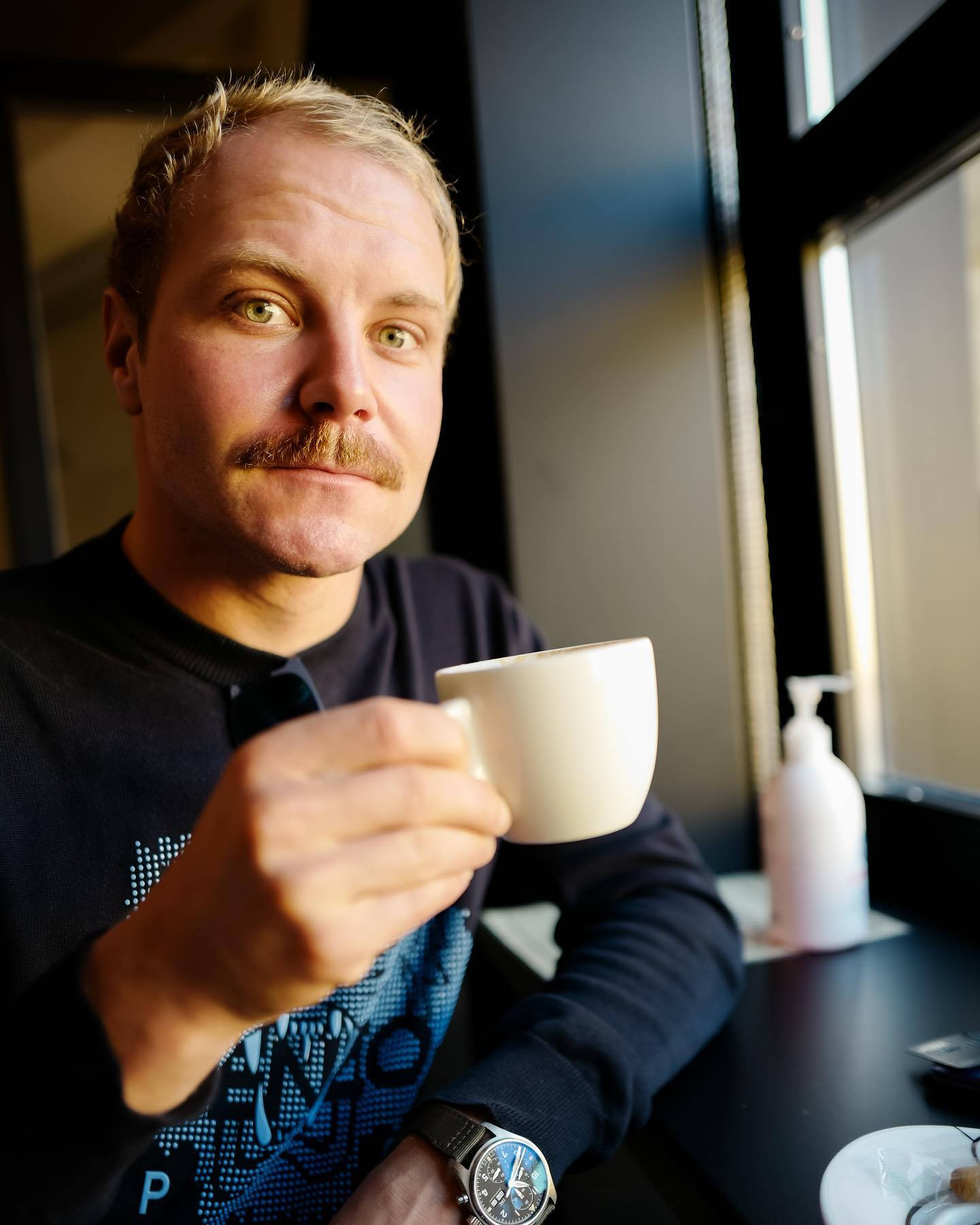 Valtteri Bottas 桌布 壁紙 — 一位留鬍鬚的男人在現代咖啡館裡拿著白色咖啡杯看著鏡頭。