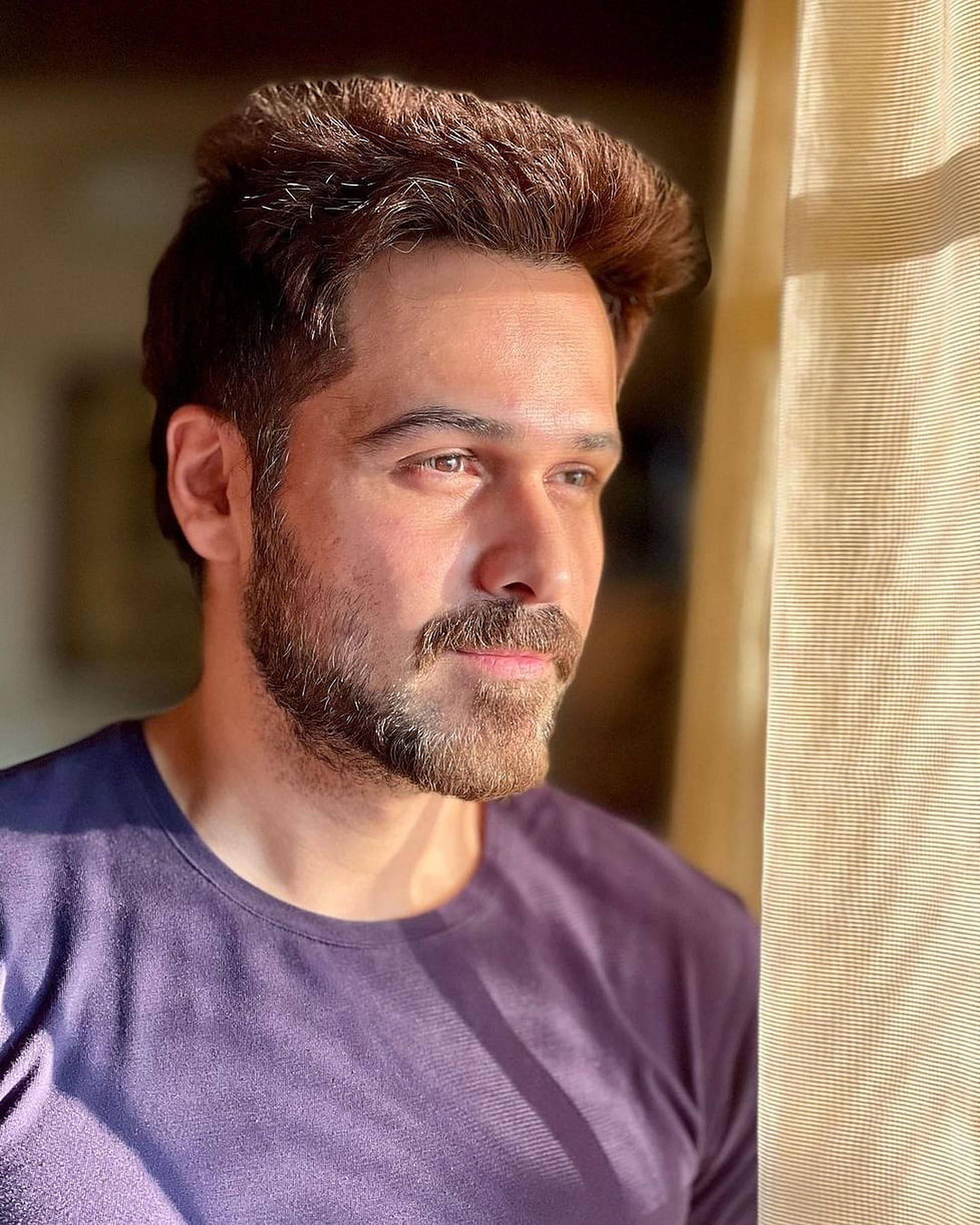 Emraan Hashmi 桌布 壁紙 — 一位留鬍鬚的男人在陽光照射的窗邊，金色窗簾框著，若有所思地凝視著。