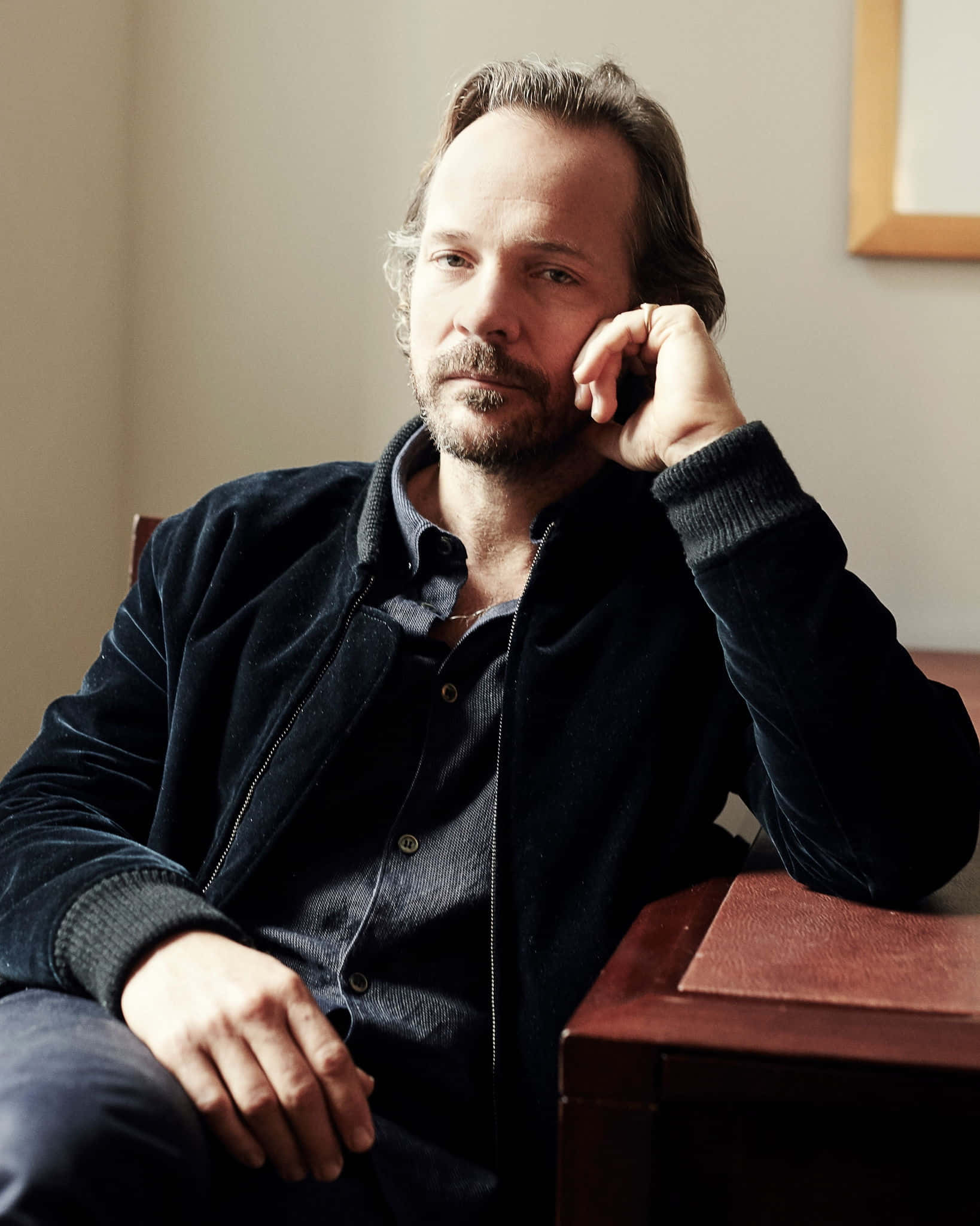 Peter Sarsgaard 桌布 壁紙 — 一位留鬍鬚的男人坐在椅子上，手托著臉頰，沉思著，身穿深色天鵝絨外套。