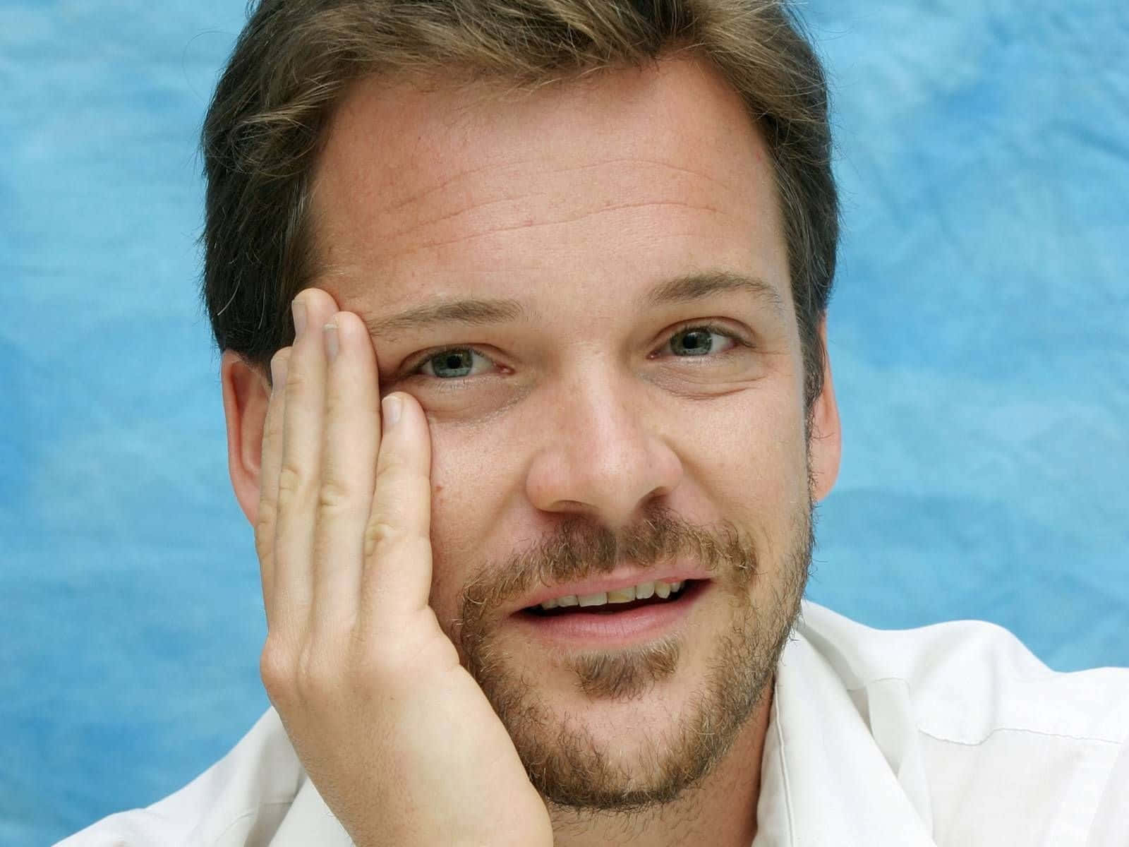 Peter Sarsgaard 桌布 壁紙 — 一位留鬍鬚的男人將手放在臉頰上，在帶有紋理的藍色背景前溫暖地微笑。