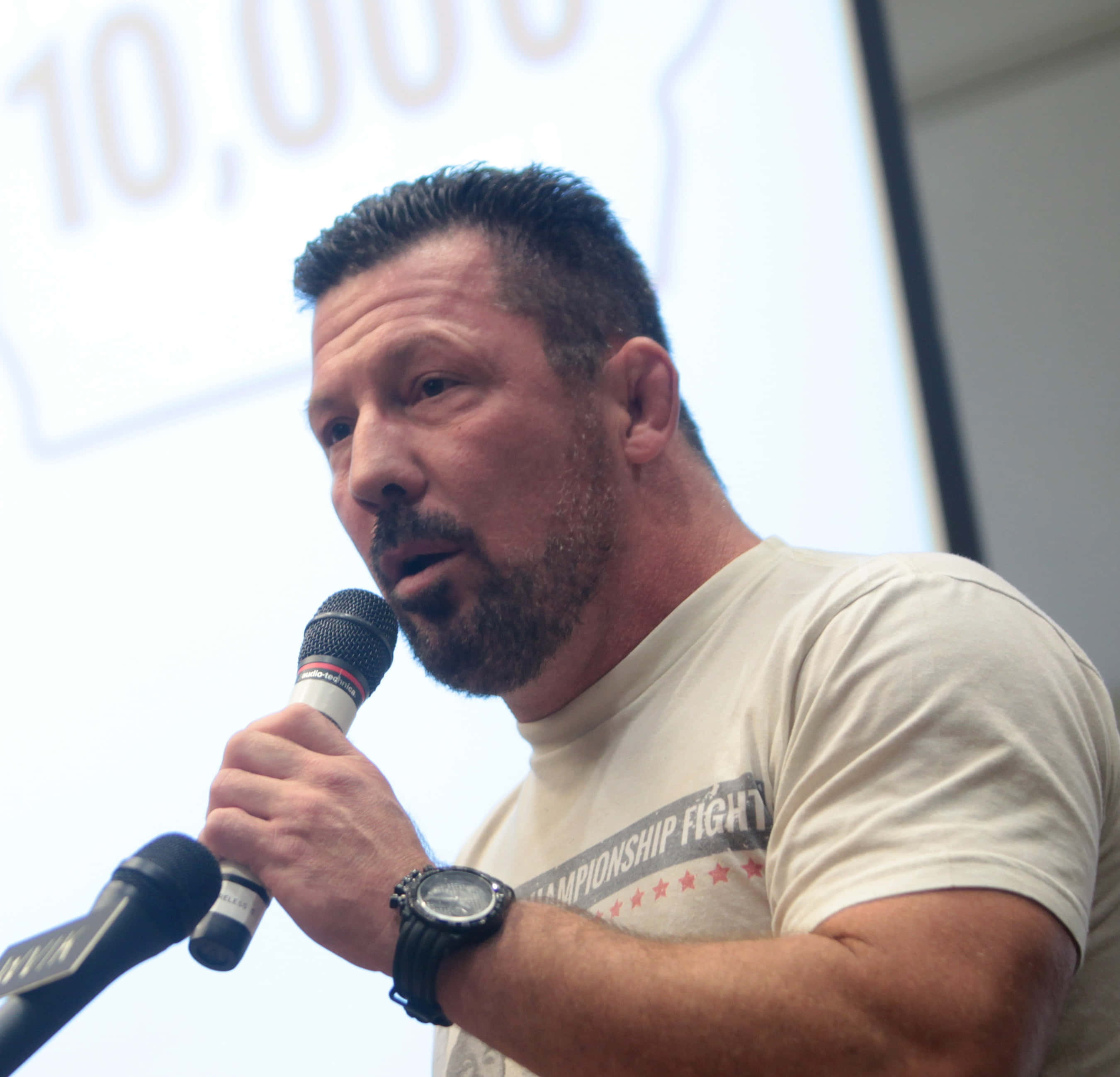 Coach 桌布 壁紙 — 一位留鬍鬚的男人對著麥克風說話，身穿冠軍爭奪戰T恤和黑色手錶。