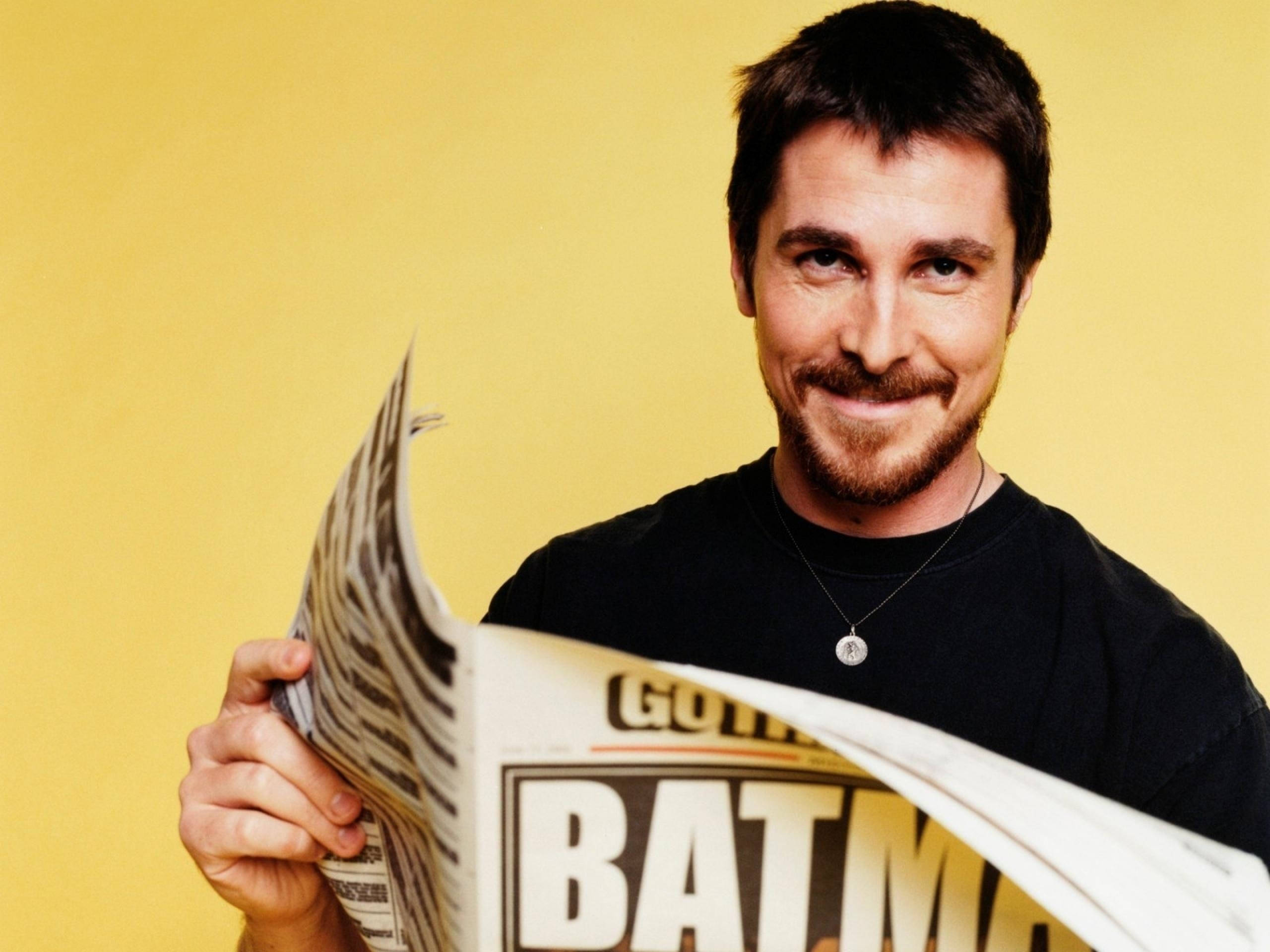 Christian Bale 桌布 壁紙 — 一位留鬍鬚的男人微笑著舉起一份報紙，上面有「BATMAN」字樣，背景為純黃色。