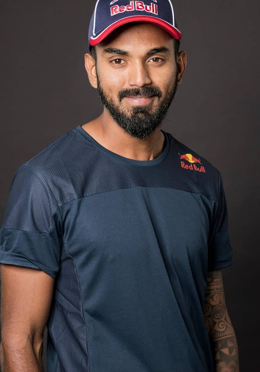 Red Bull 桌布 壁紙 — 一位留鬍鬚的男人戴著Red Bull帽和運動衫，在深色攝影棚背景下自信微笑。
