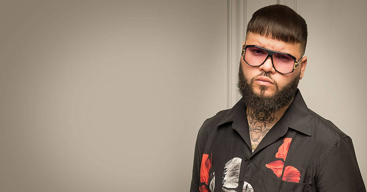 farruko 桌布 壁紙 — 一位留鬍鬚的男人戴著大膽的粉紅色太陽眼鏡，自信地站在純米色牆前面，穿著一件有紅色花卉圖案的黑色襯衫。