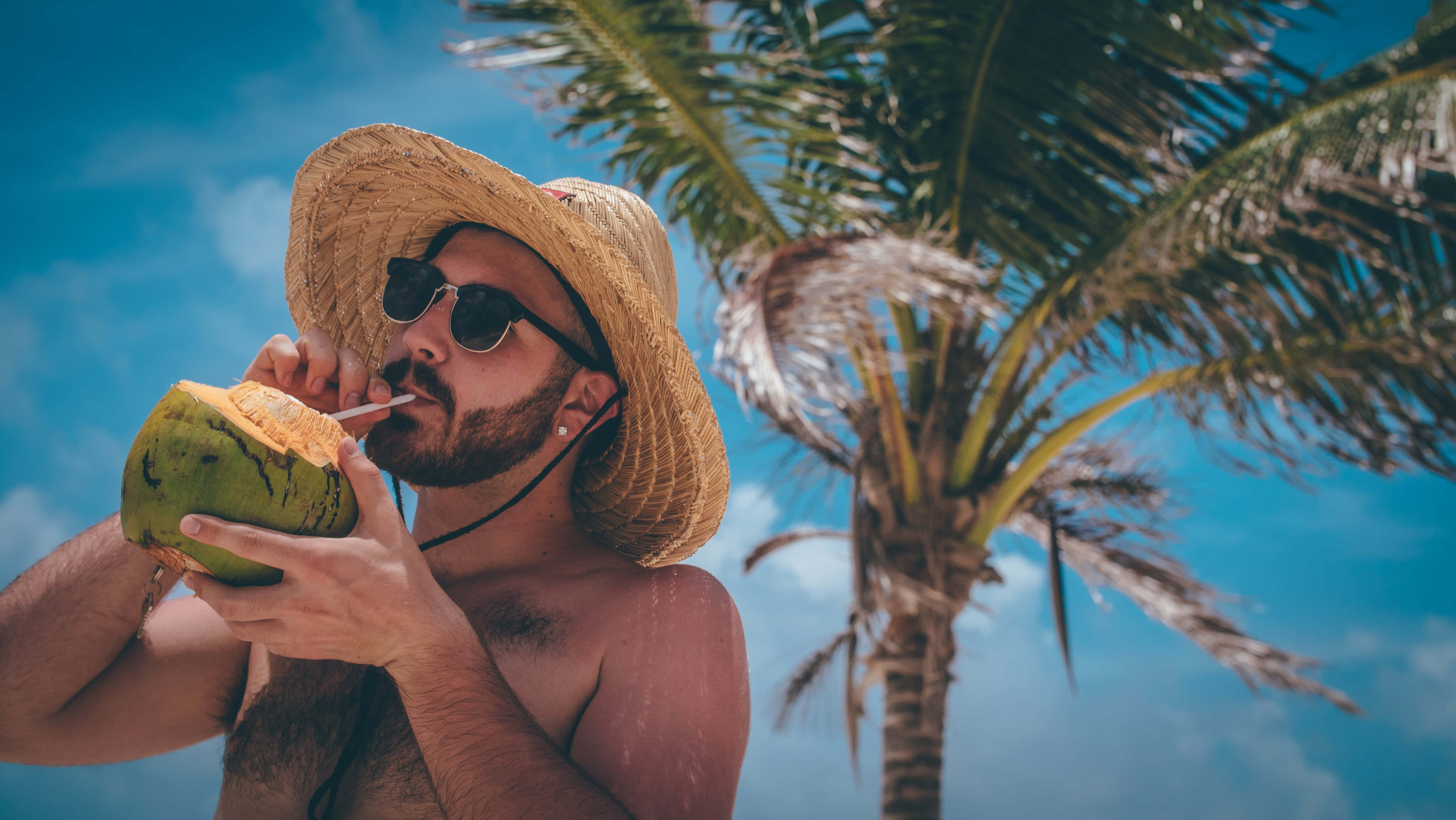 Summer Vacation 桌布 壁紙 — 一位留鬍鬚的男人戴著草帽和太陽眼鏡，在棕櫚樹下喝椰子水，背景是明亮的藍天。