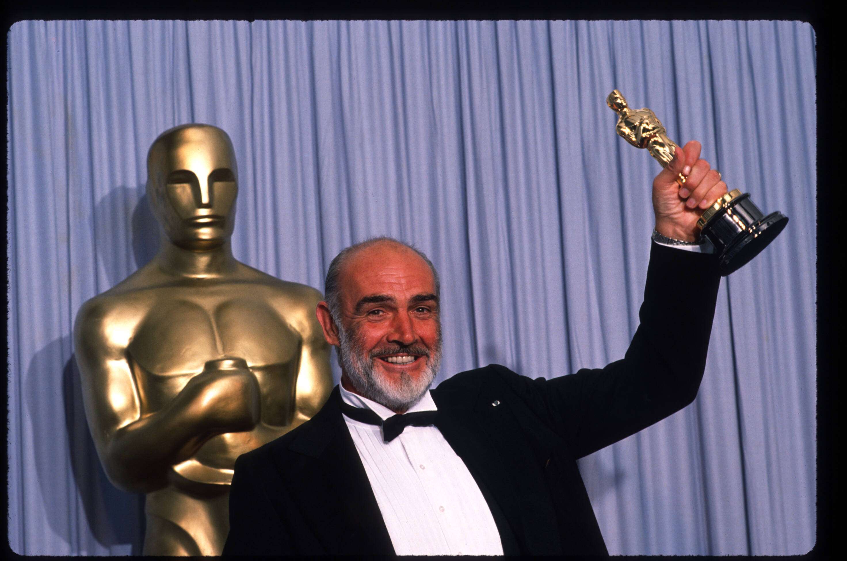 Sean Connery 桌布 壁紙 — 一位留鬍鬚的男人穿著燕尾服微笑，手持奧斯卡獎座，旁邊有一個巨大的金色奧斯卡獎座，背景是藍色窗簾。