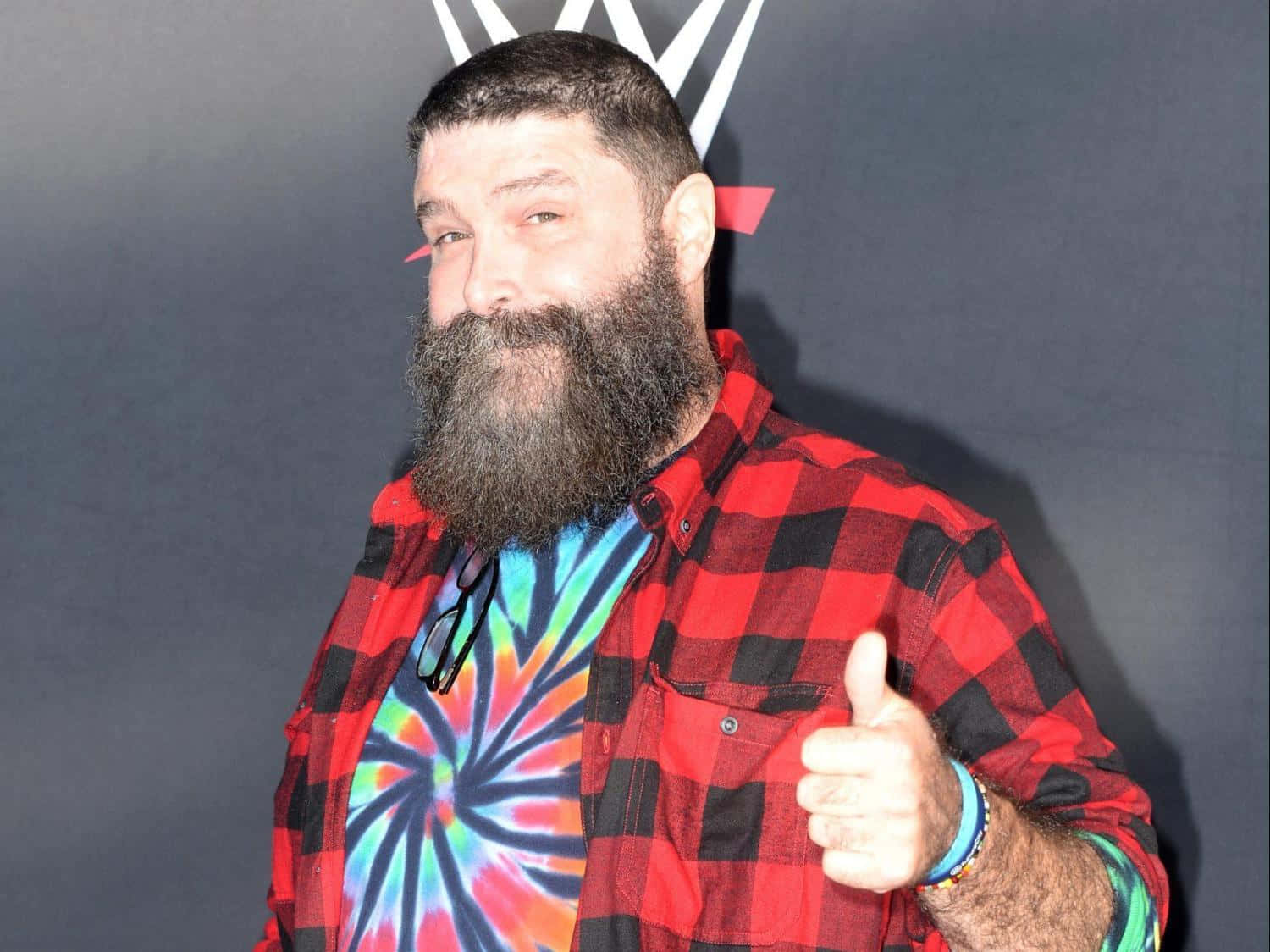 WWE 桌布 壁紙 — 一位留鬍鬚的男人穿著紅色格紋襯衫內搭紮染T恤，在黑暗背景前豎起大拇指，背景有WWE標誌。