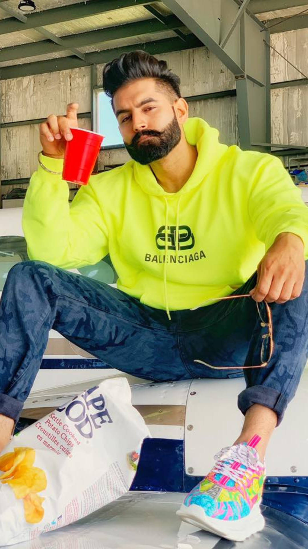 Indian 桌布 壁紙 — 一位留鬍鬚的男人穿著螢光黃色連帽衫，隨意地坐著，手持紅色杯子，旁邊有一袋薯片。
